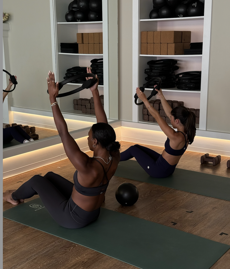 Two women on mats doing Pilates with rings in a workout room with wooden floor and shelves of equipment.