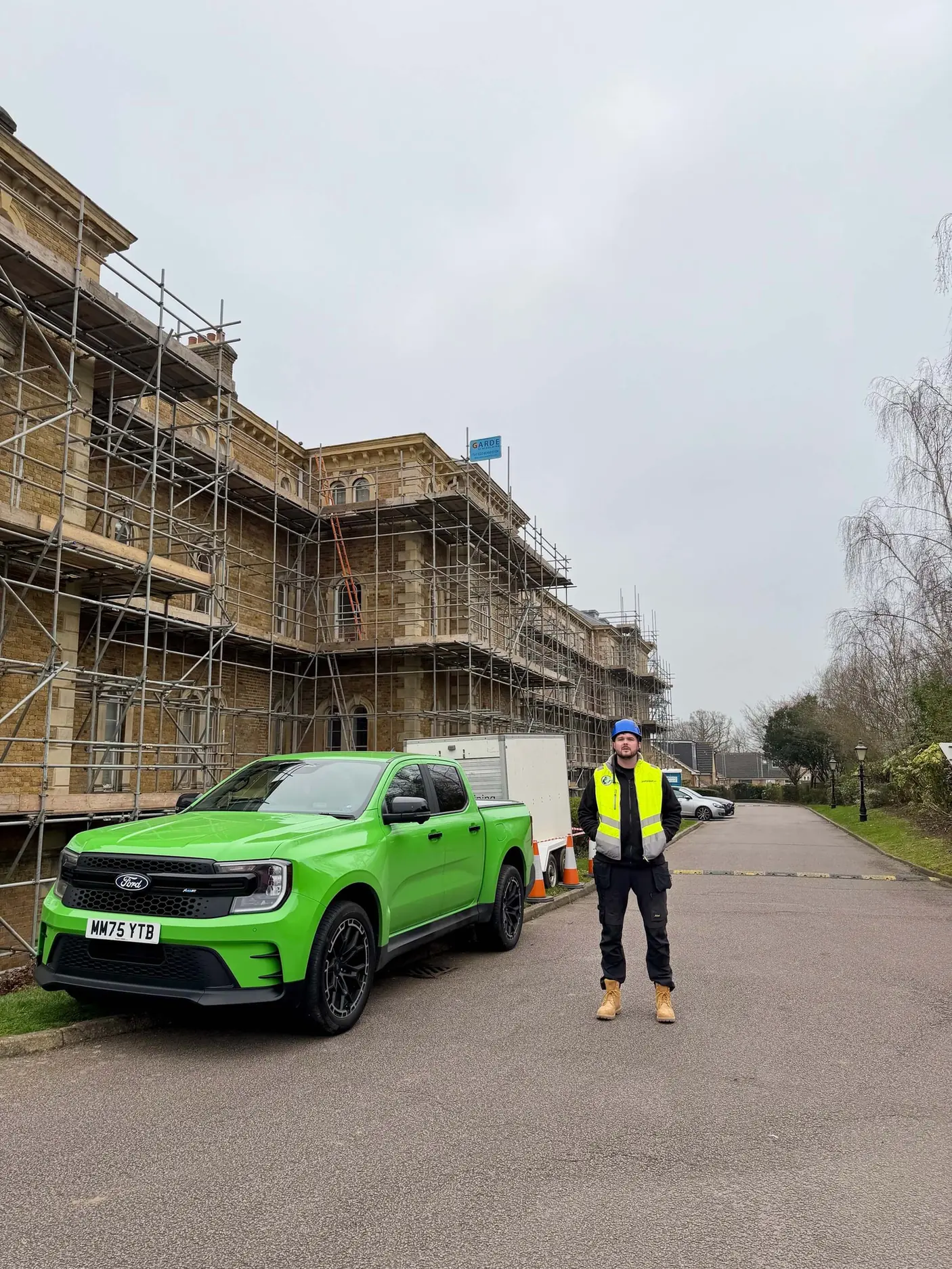 Man in a blue hard hat and yellow vest stands on road by bright green Ford truck near building with scaffolding.