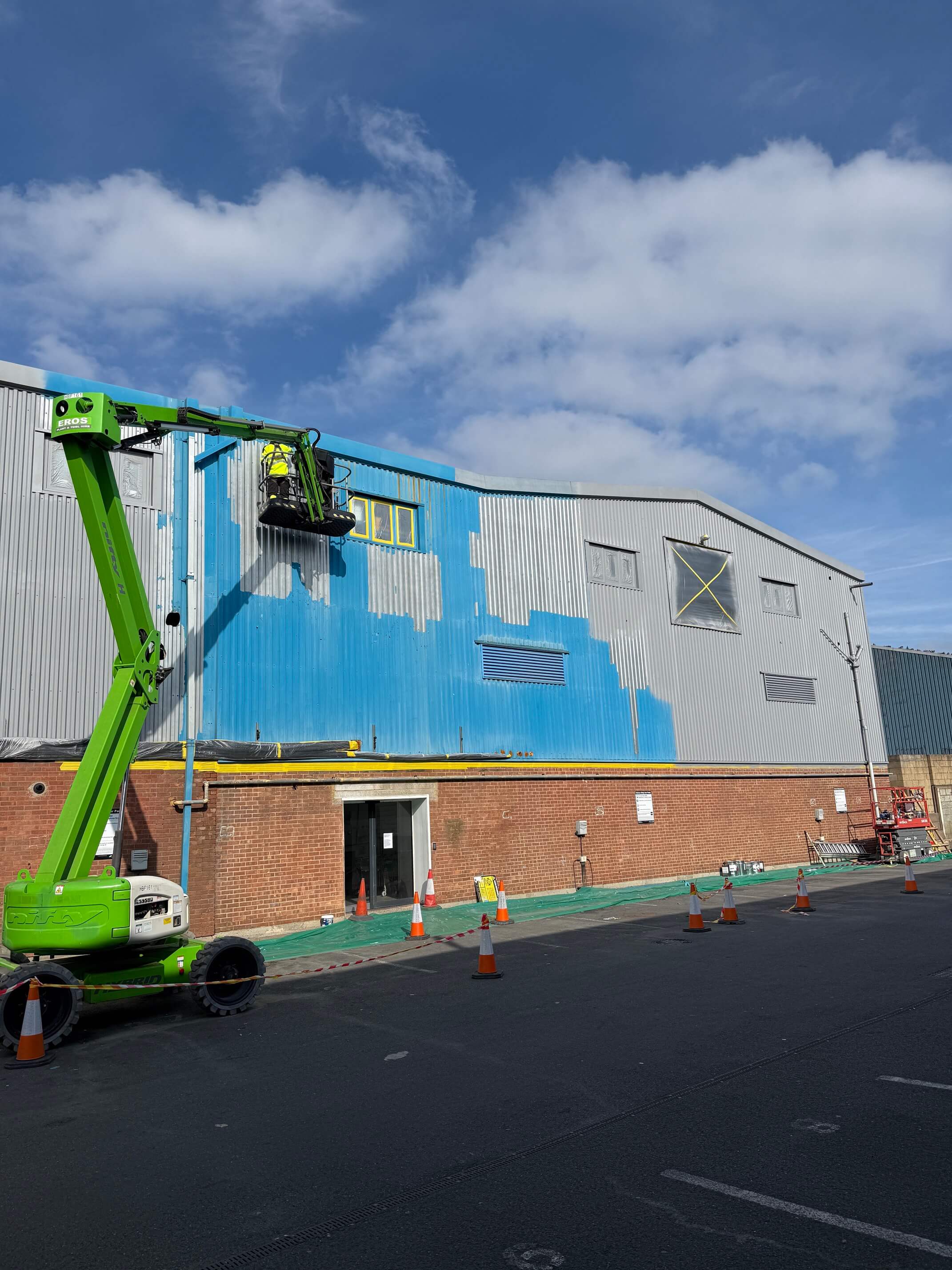 Worker using green boom lift to spray paint blue on upper metal wall of industrial building.
