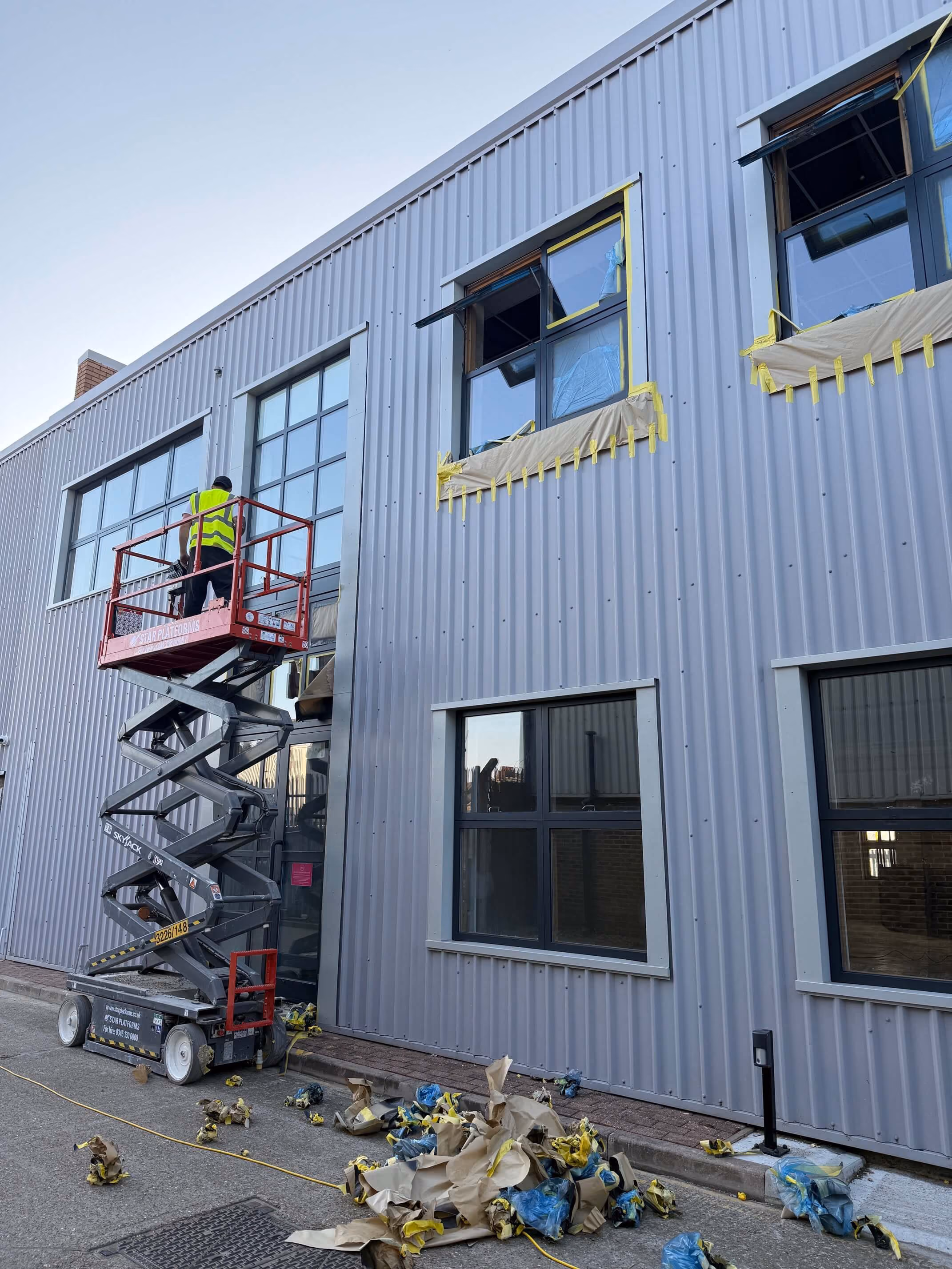 Worker in high-visibility vest on scissor lift installing or cleaning windows on metal warehouse facade.