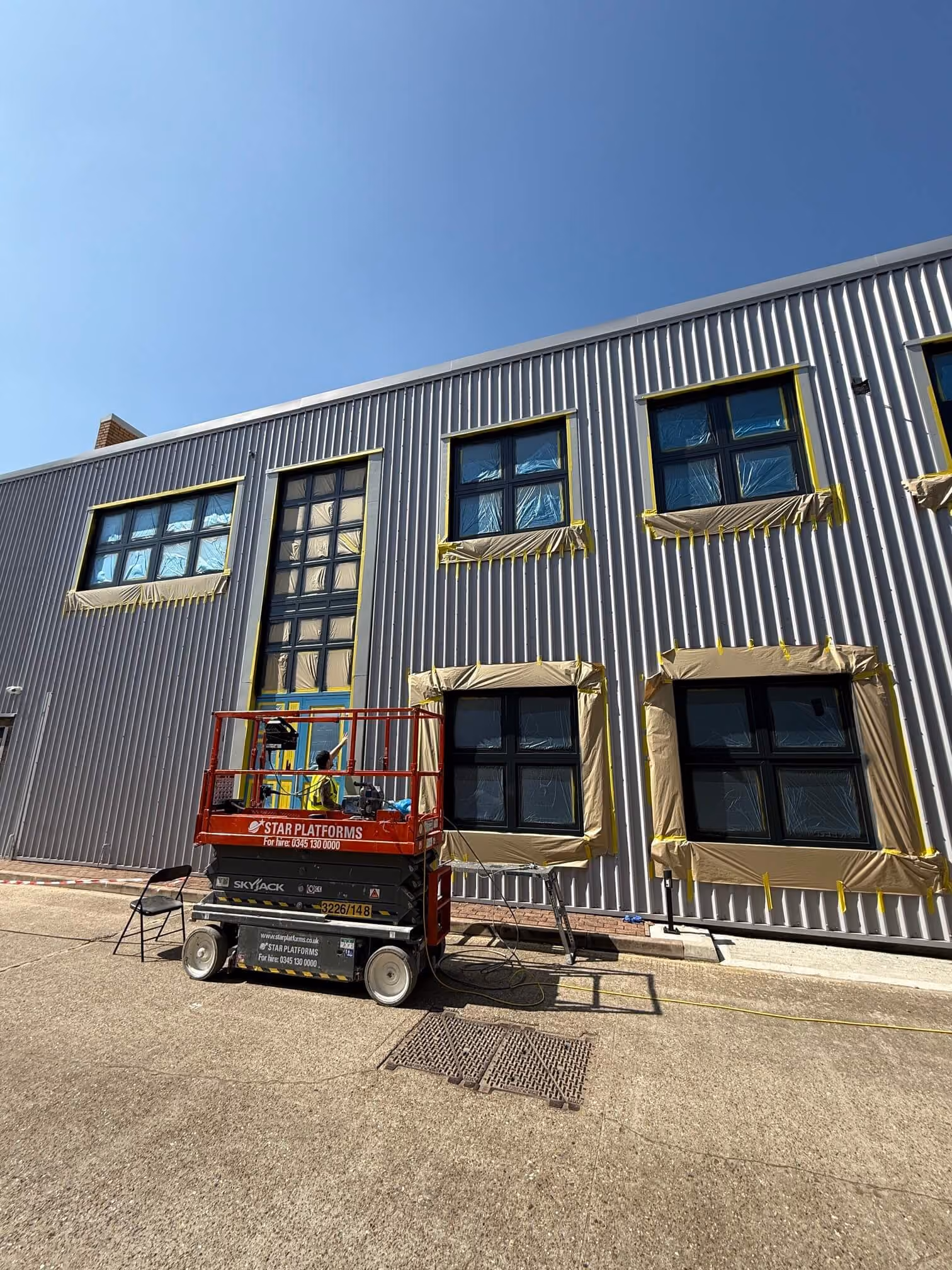 Worker on a red scissor lift preparing a building's windows and door for painting with protective sheeting.