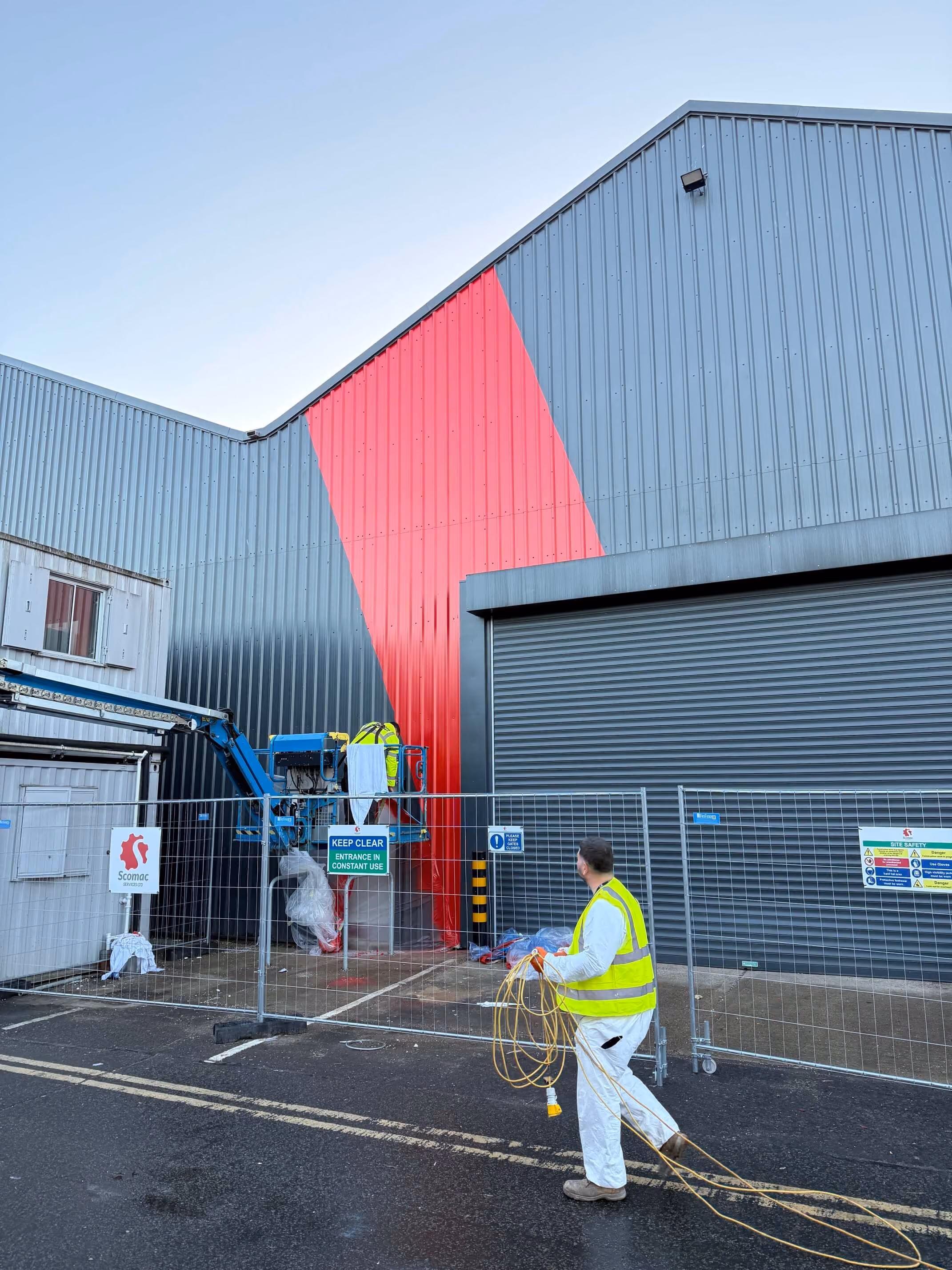 Worker in high-visibility vest holding extension cords near building with red diagonal stripe on metal cladding.
