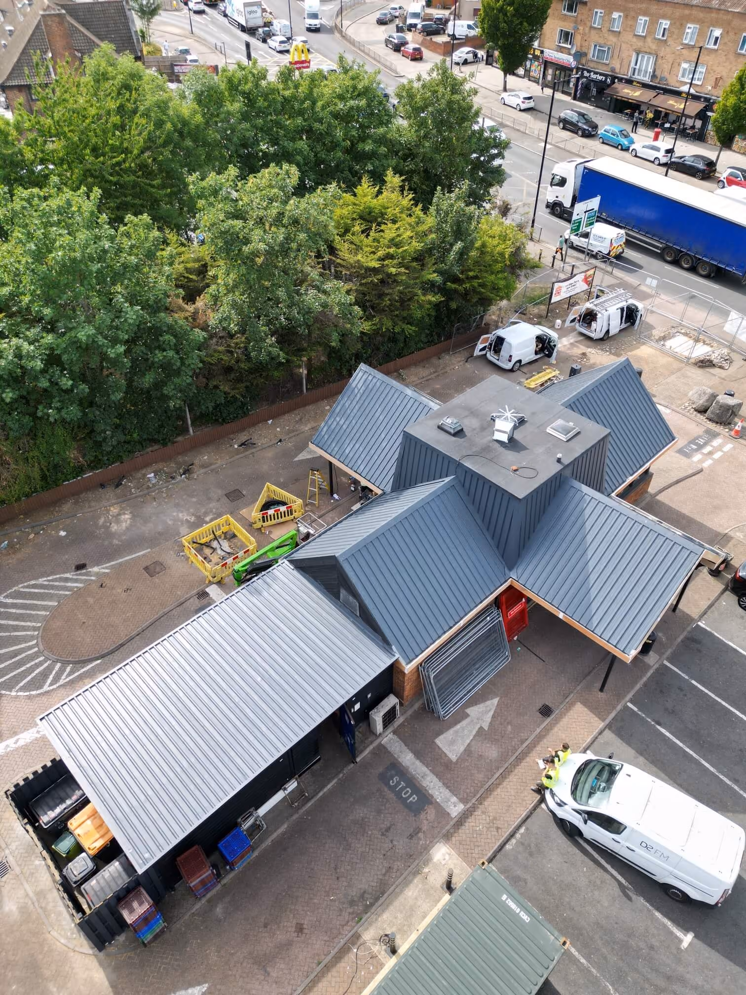 Aerial view of a small building with gray metal roofs in a parking area near a busy road and trees.