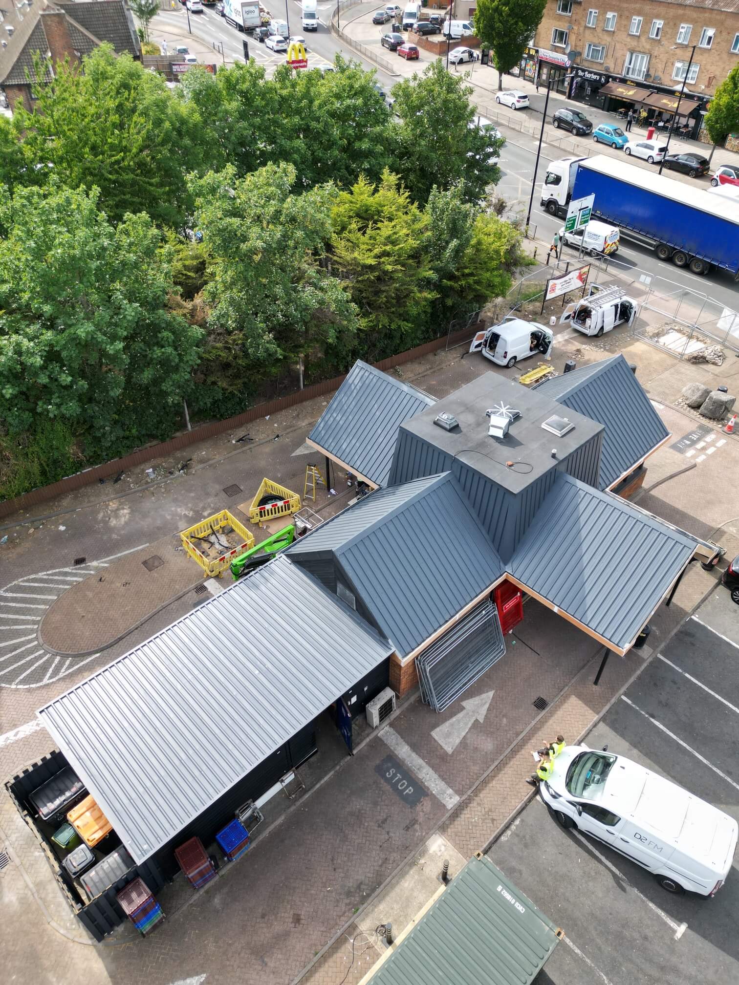 Aerial view of a small building with gray metal roofs in a parking area near a busy road and trees.