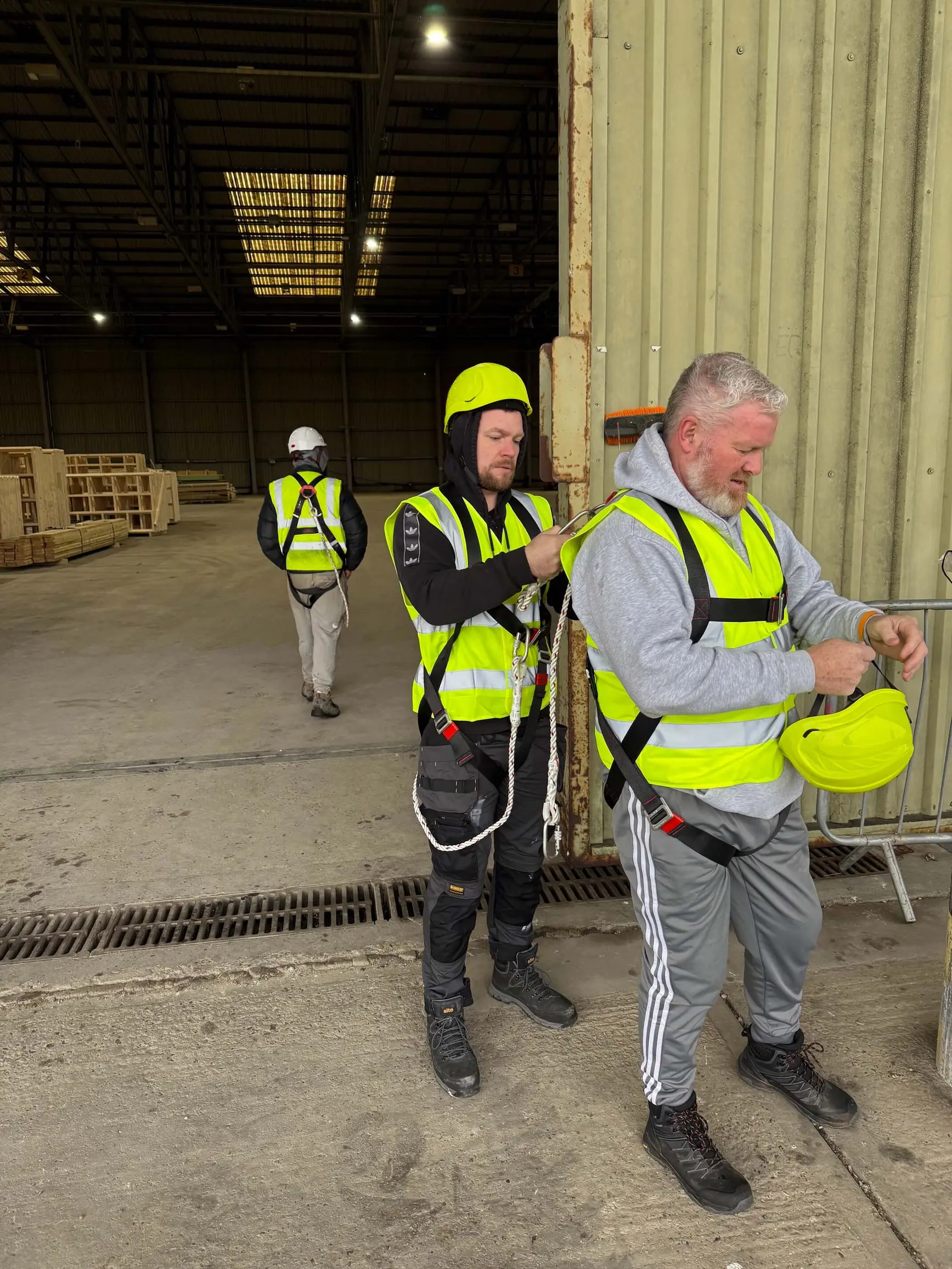 Three workers wearing high-visibility vests and safety harnesses inside a warehouse.