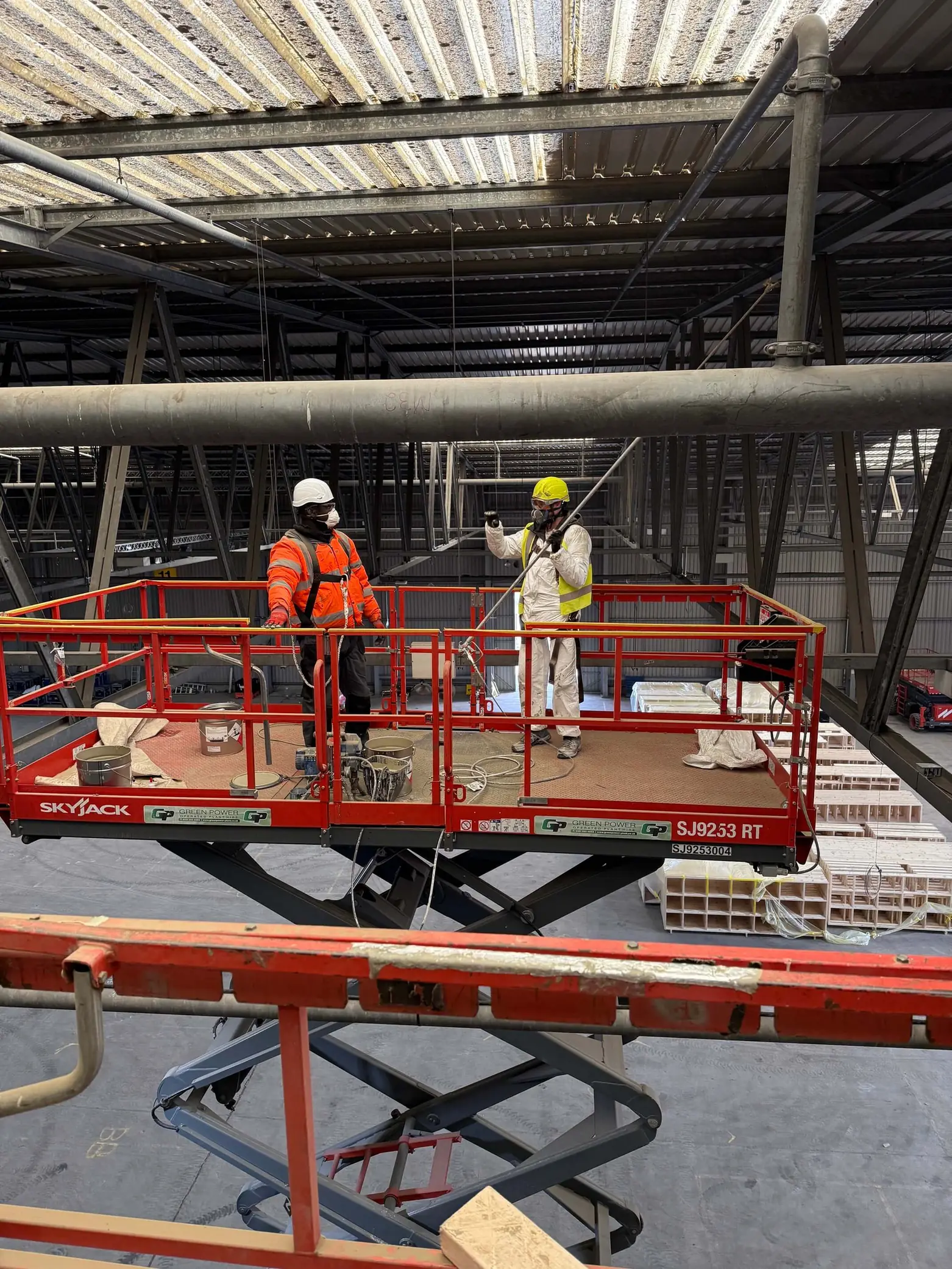 Two workers wearing safety gear stand on a red scissor lift inside a large industrial warehouse.