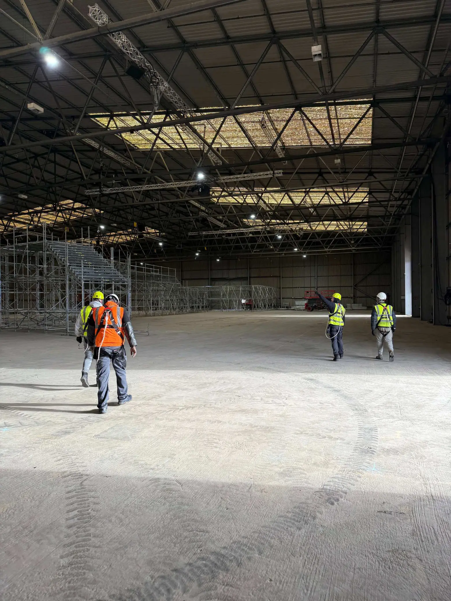 Four workers in safety gear walk inside a large warehouse with metal scaffolding along the left wall.