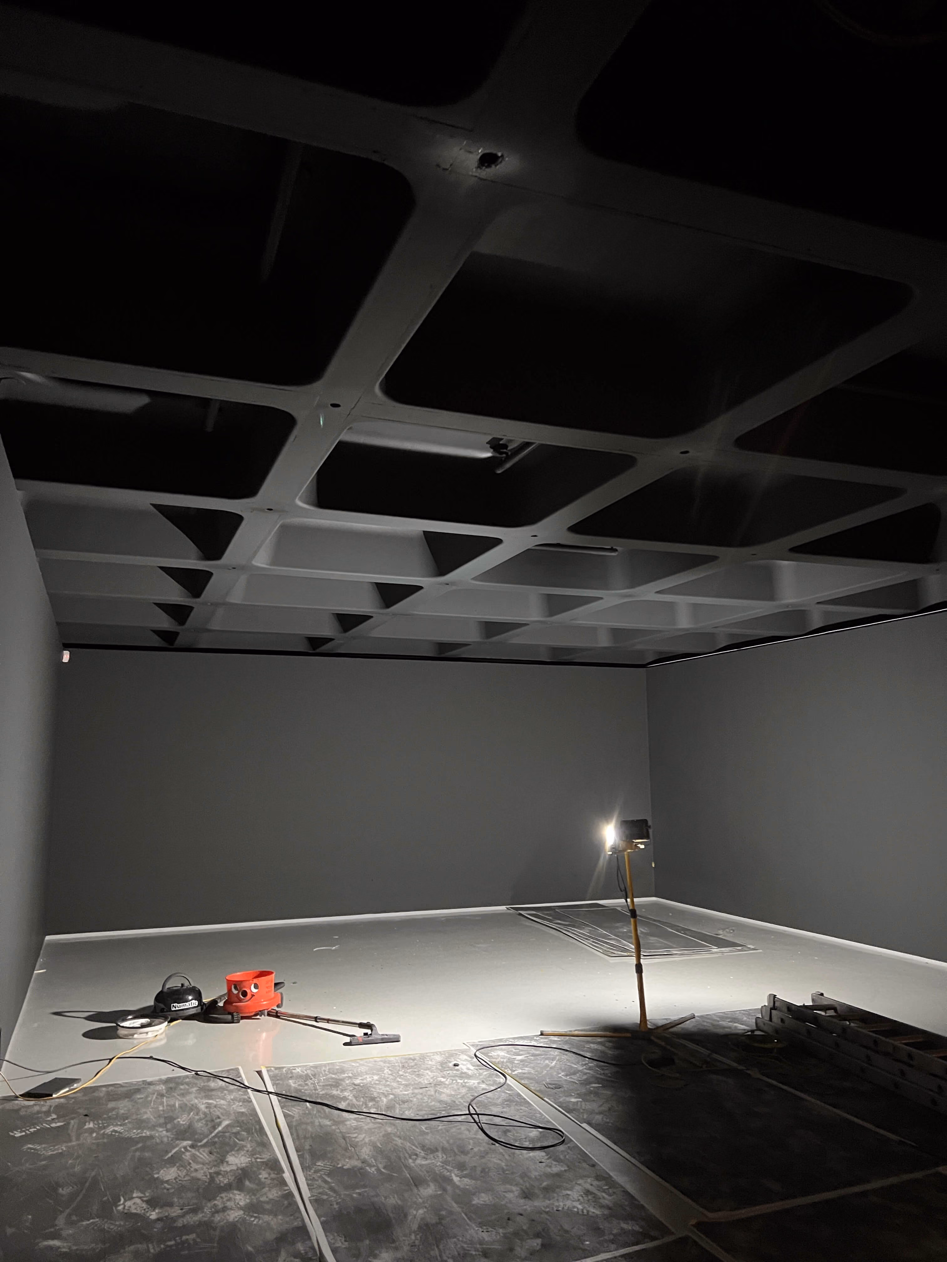 Dimly lit empty room with dark walls, ceiling grid, and cleaning equipment on the floor.