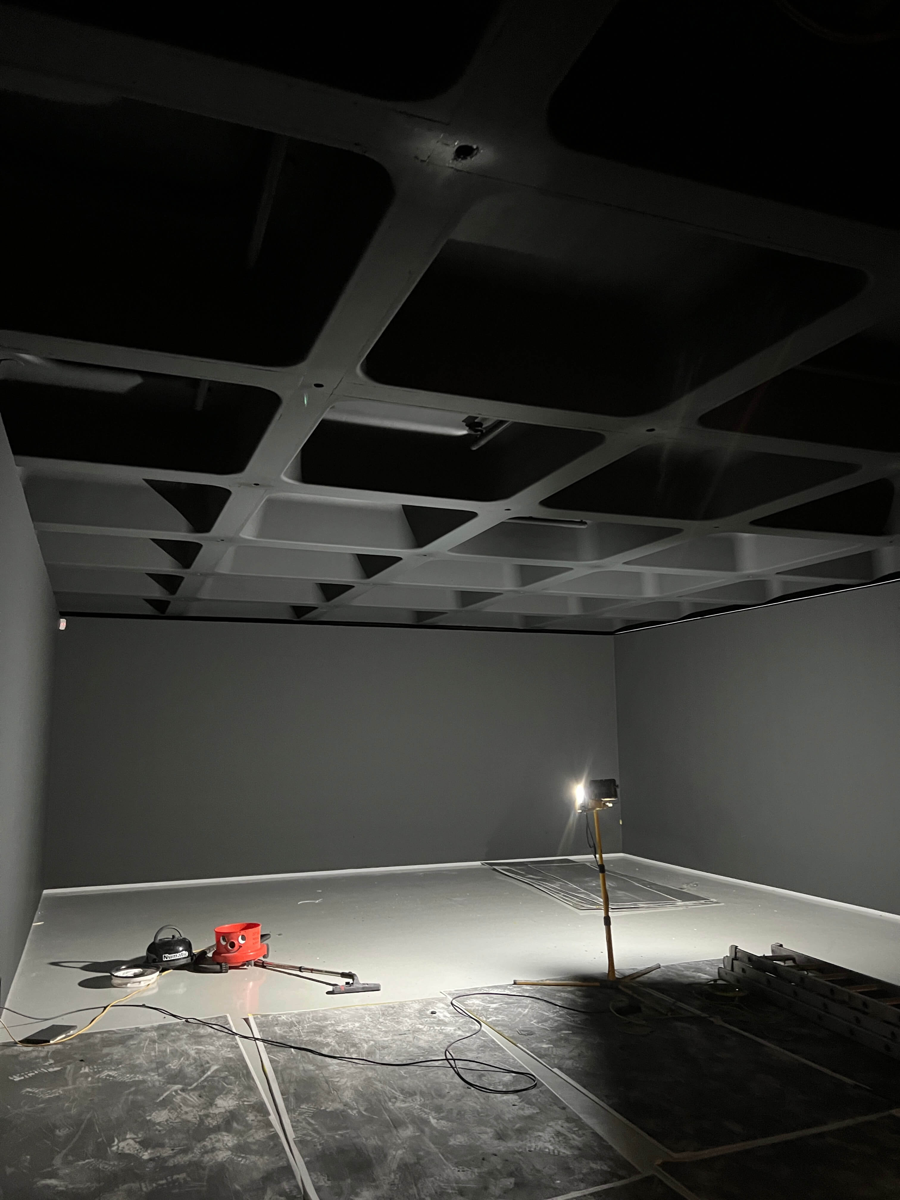 Dimly lit empty room with dark walls, ceiling grid, and cleaning equipment on the floor.
