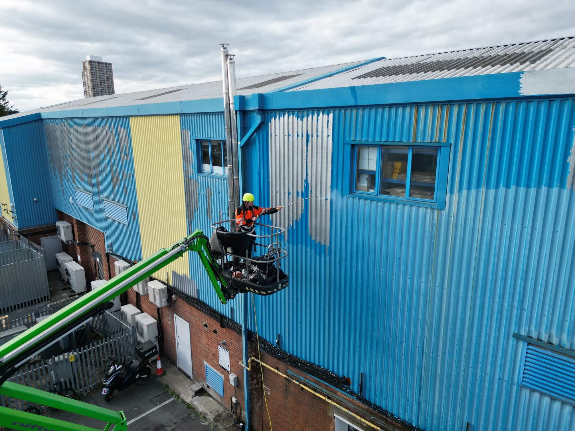 Worker in safety gear power washing a large blue industrial building exterior from an aerial lift.