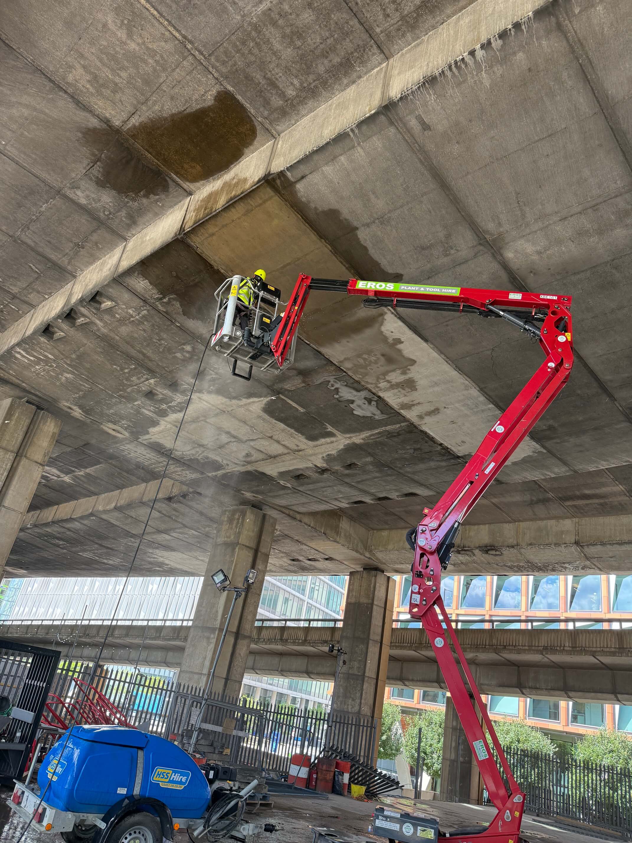 Worker uses a red aerial lift to pressure wash the underside of a concrete overpass.