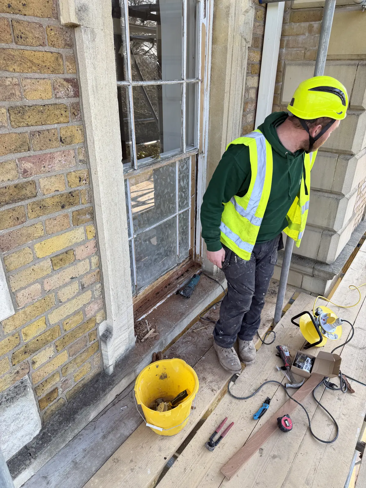 Worker in high-visibility clothing beside a timber sash window on scaffolding.