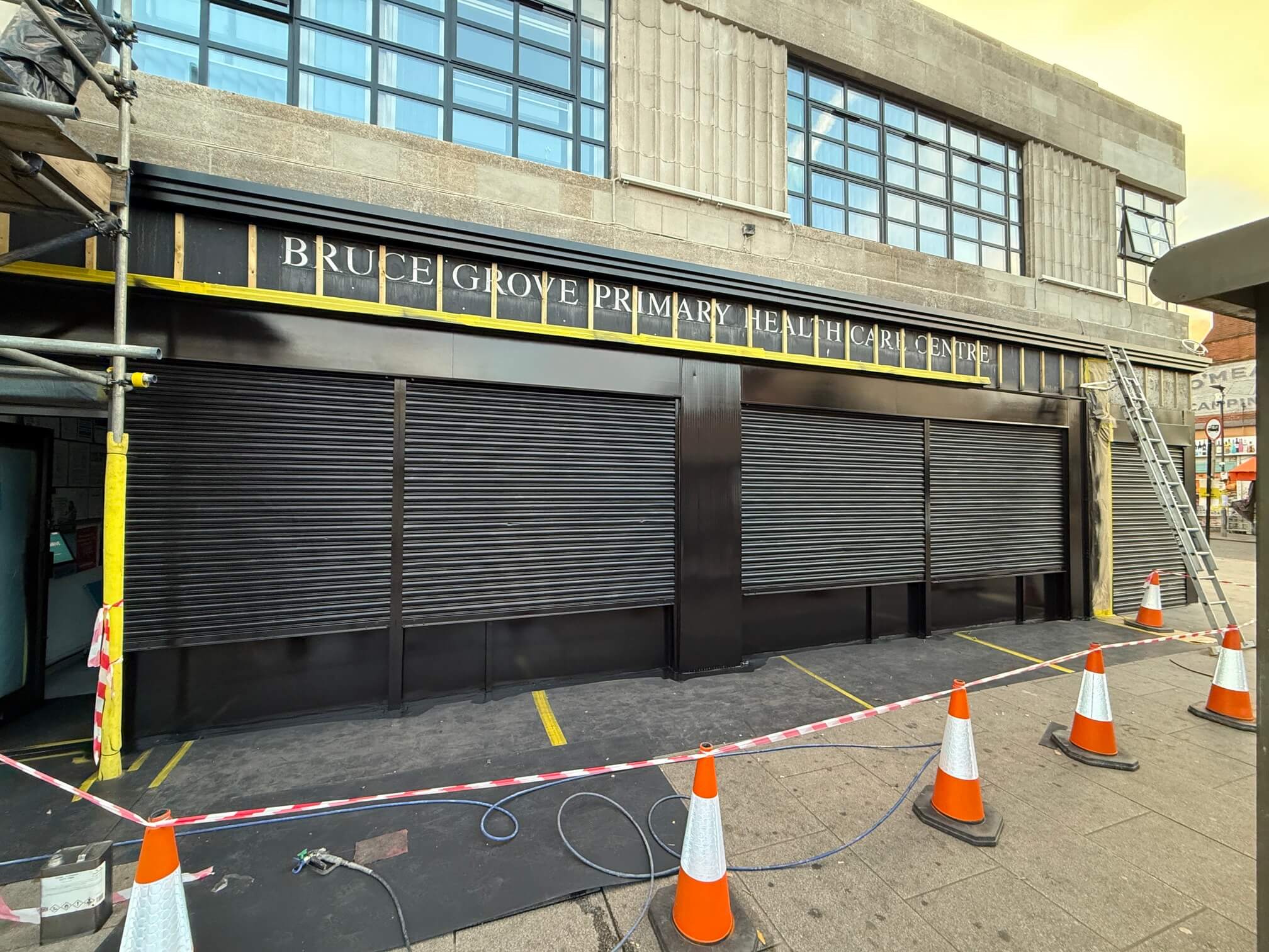 Dark-coated roller shutters outside Bruce Grove Primary Health Care Centre, with a cordoned-off work area.