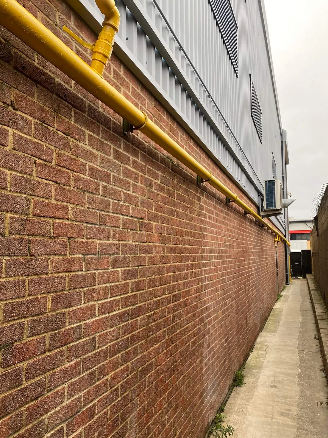 Grey metal cladding above brickwork and yellow pipework along a narrow access passage.