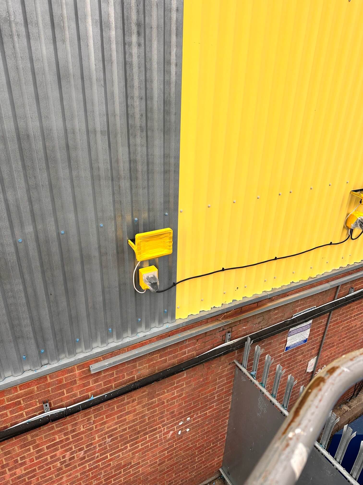 Prepared grey metal cladding beside a yellow panel, with brickwork and pipework below.