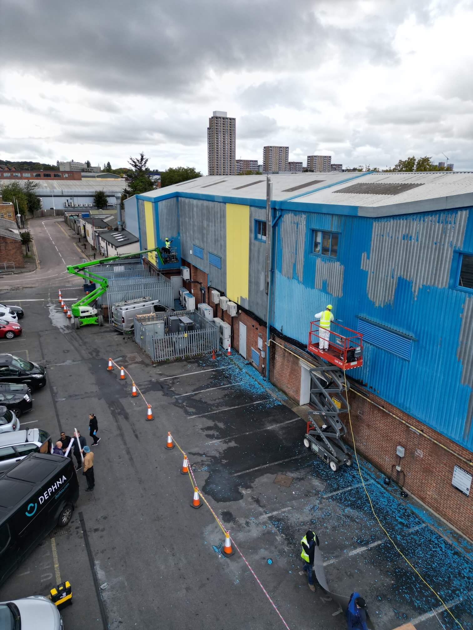 Workers on access platforms preparing blue industrial cladding beside a cordoned-off service road.