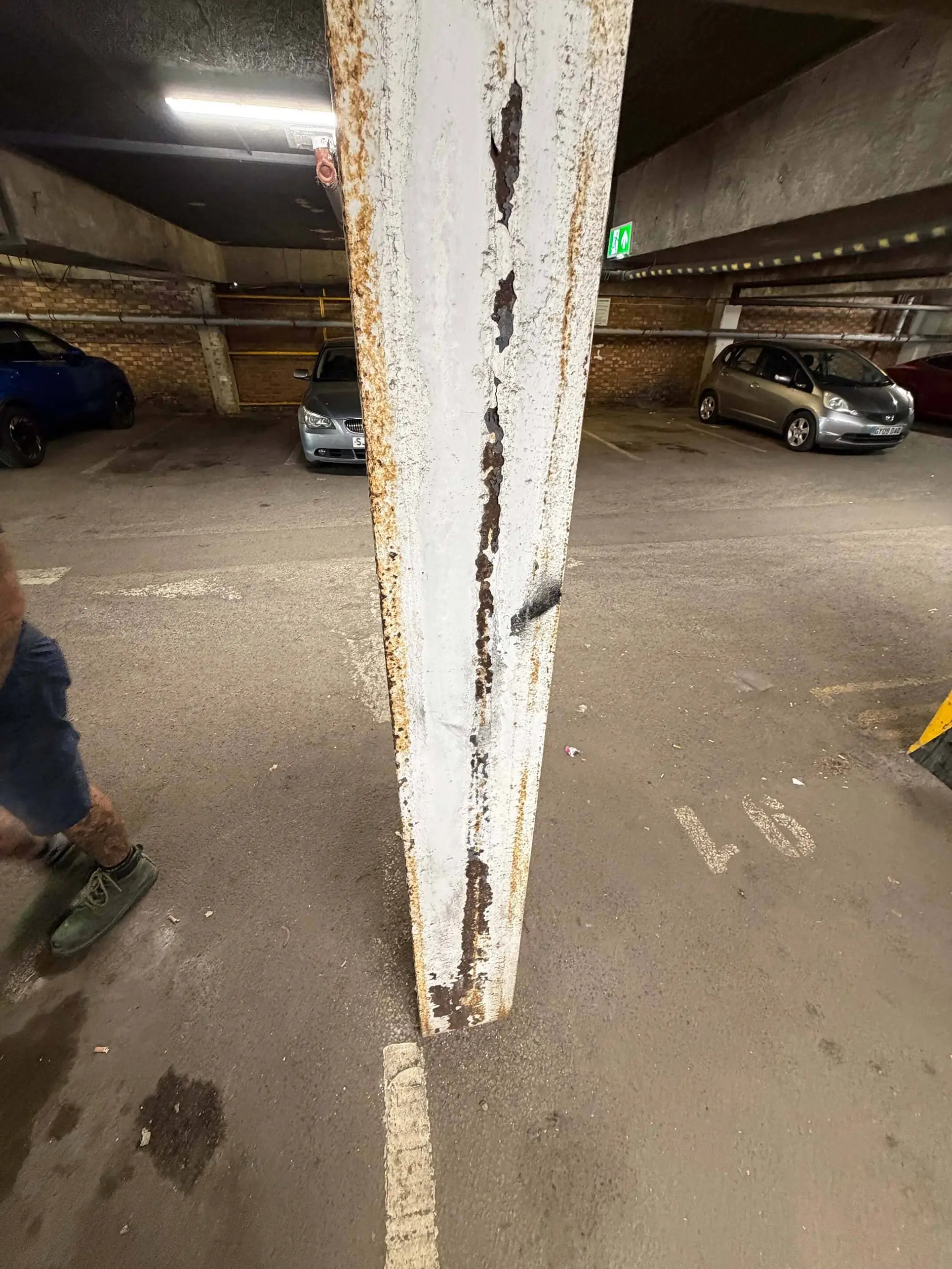 Corrosion and flaking white coating on a metal support in a car park.
