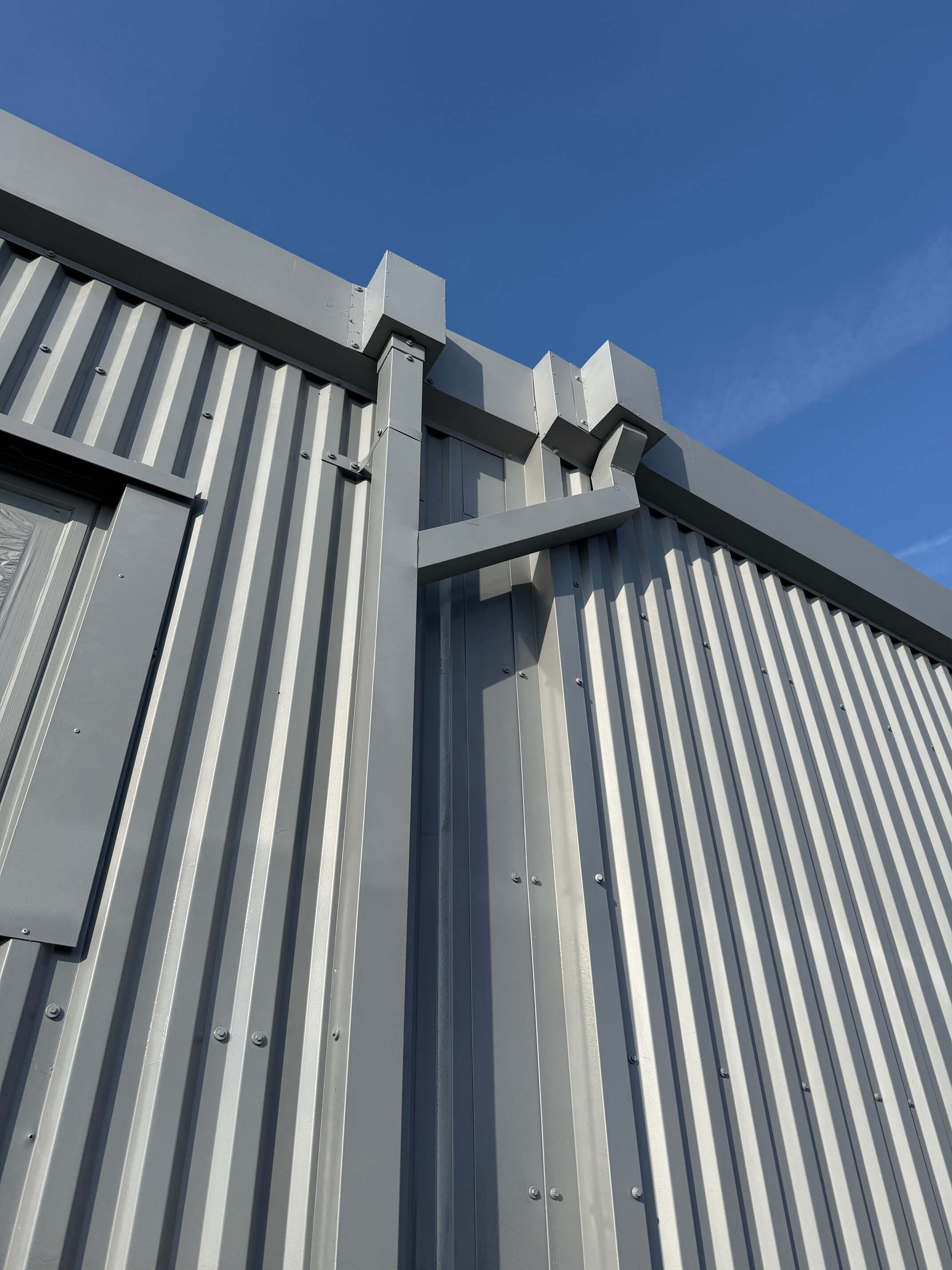 Close-up of grey profiled metal cladding and corner flashings beneath a blue sky.