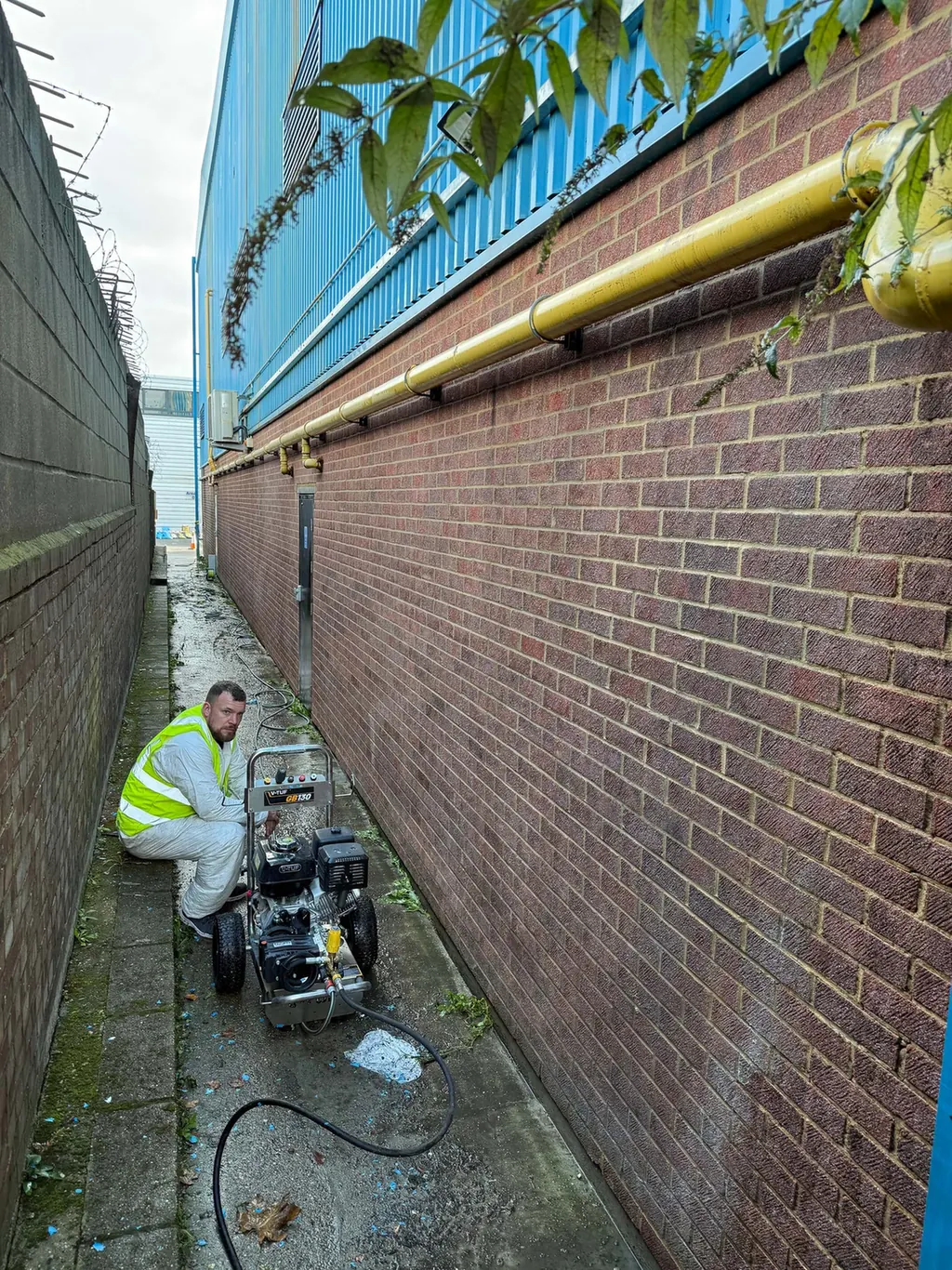 Worker beside cleaning equipment in a narrow passage alongside a brick and metal-clad building.
