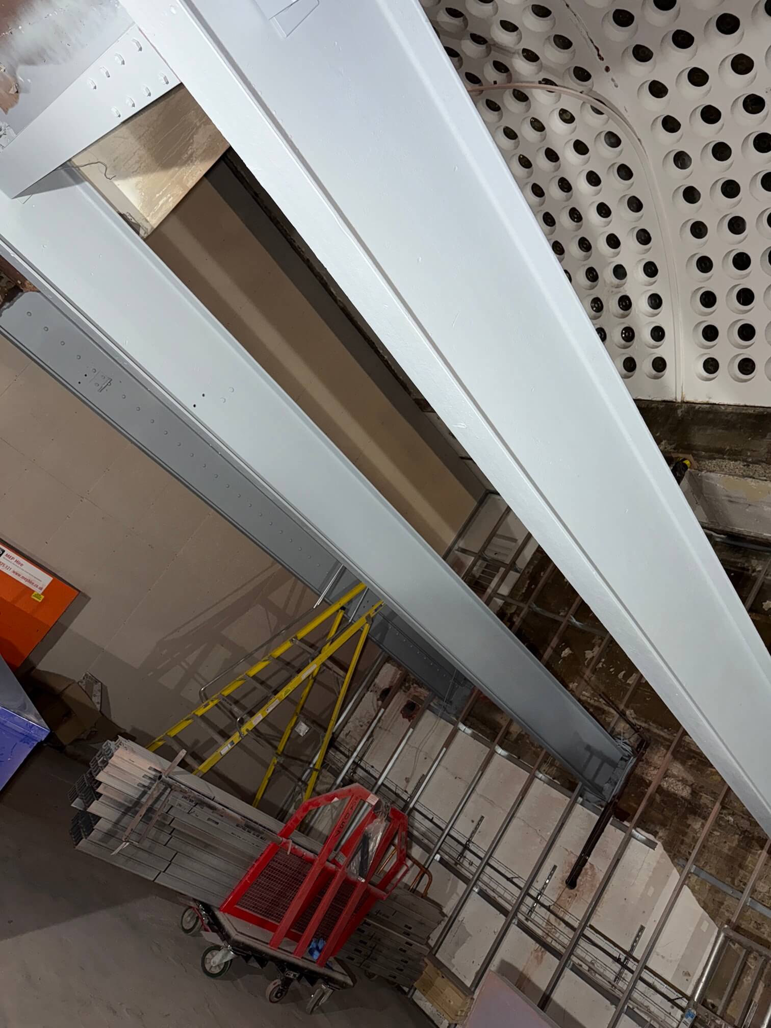 Light-coloured structural steel beams beside a perforated ceiling and access equipment.