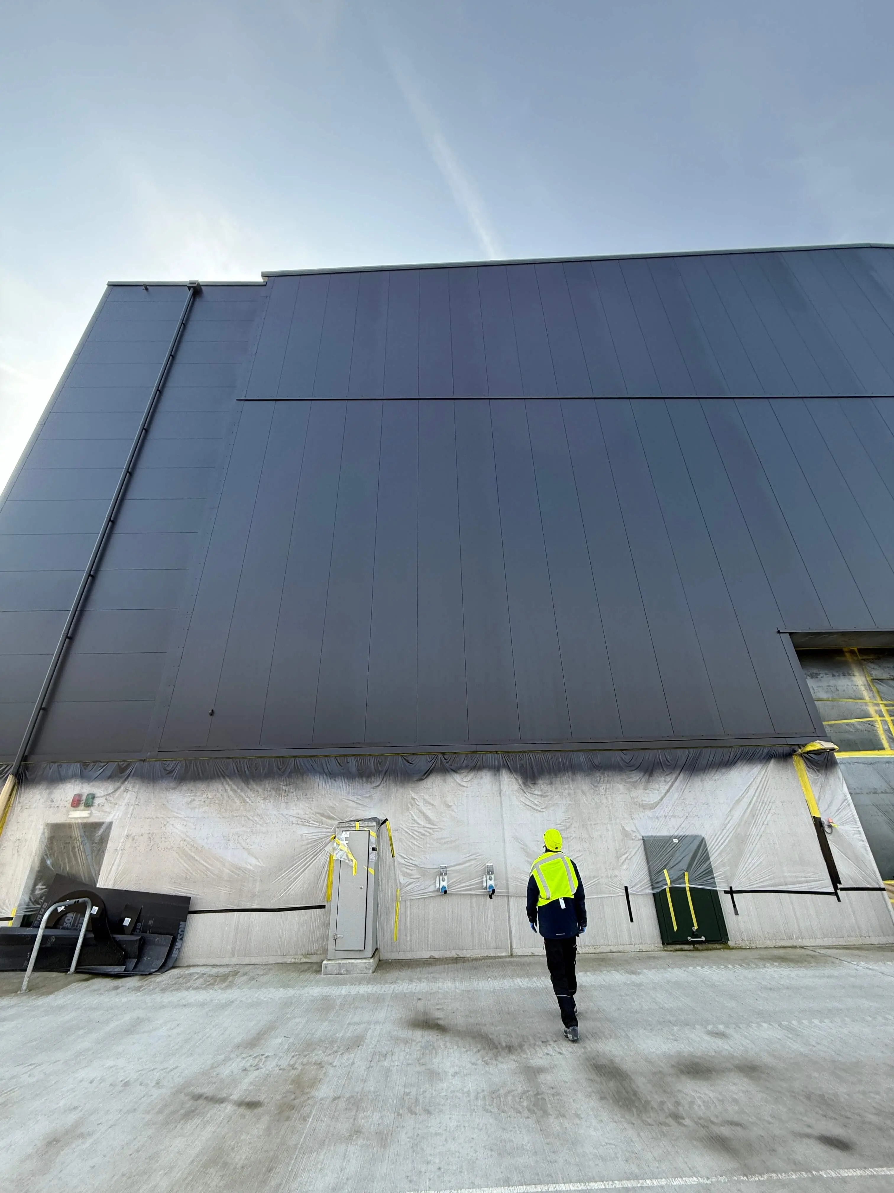 Worker in front of a dark-coated industrial facade with protected surfaces at ground level.
