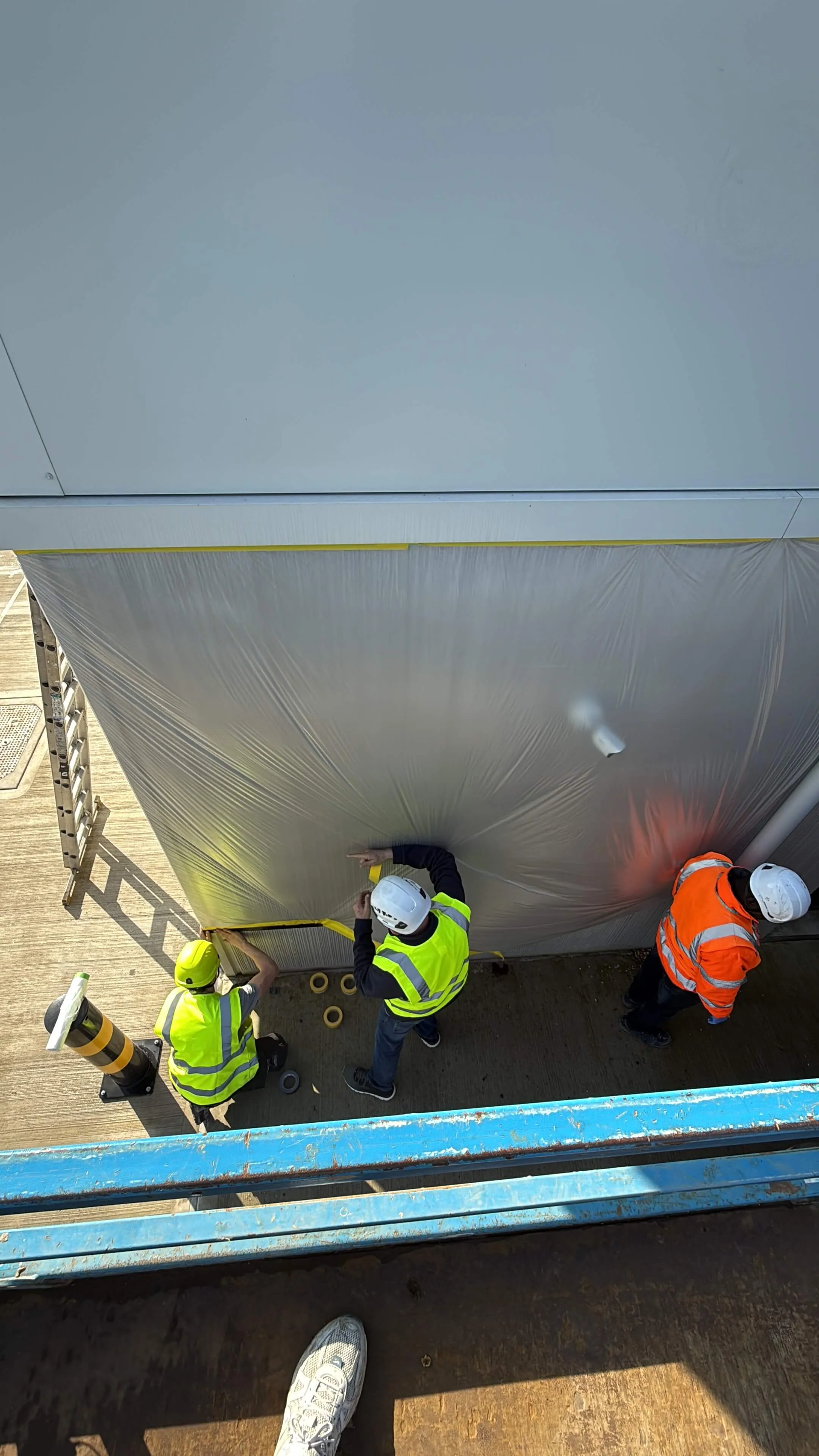 Workers beside a masked lower wall beneath newly coated industrial cladding.