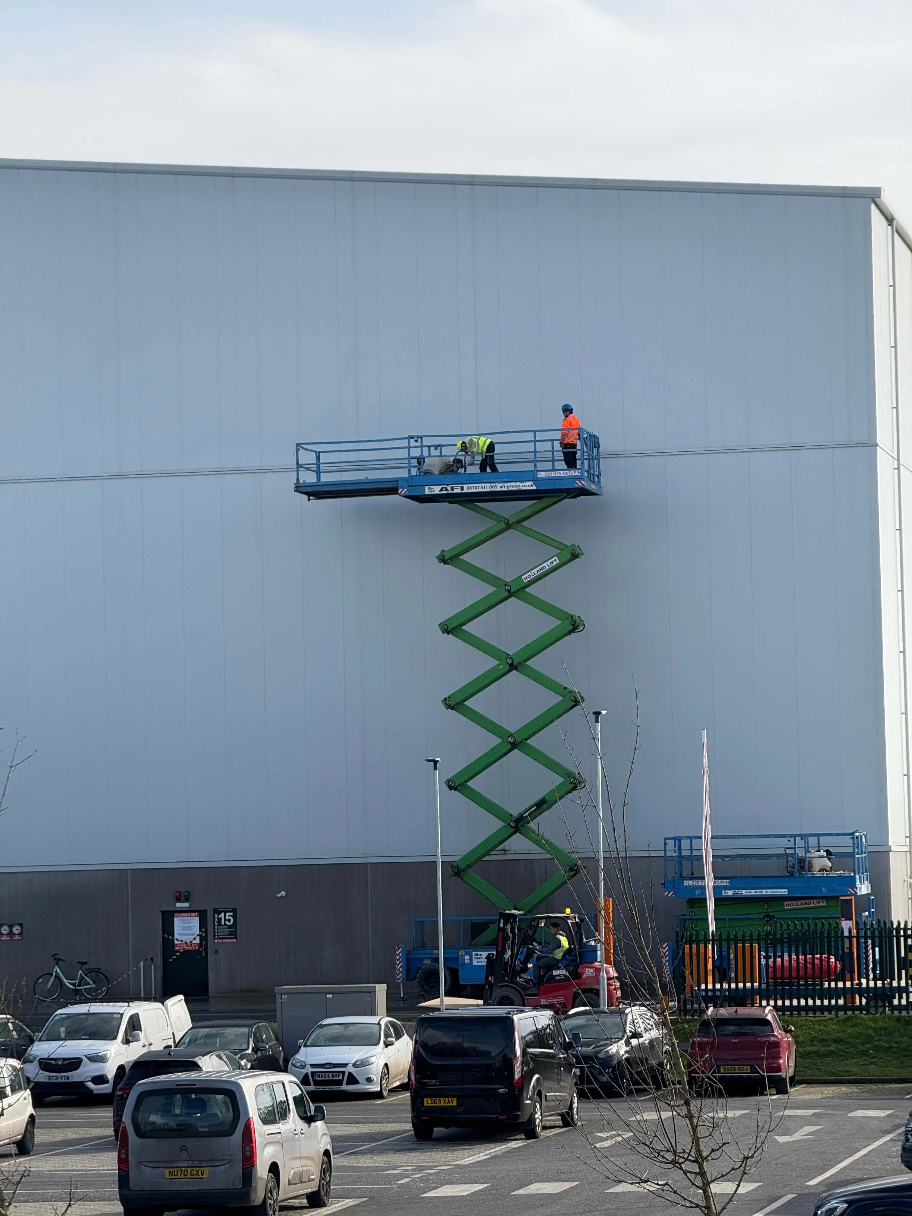 Workers on a raised scissor lift beside a large light-coloured industrial facade.