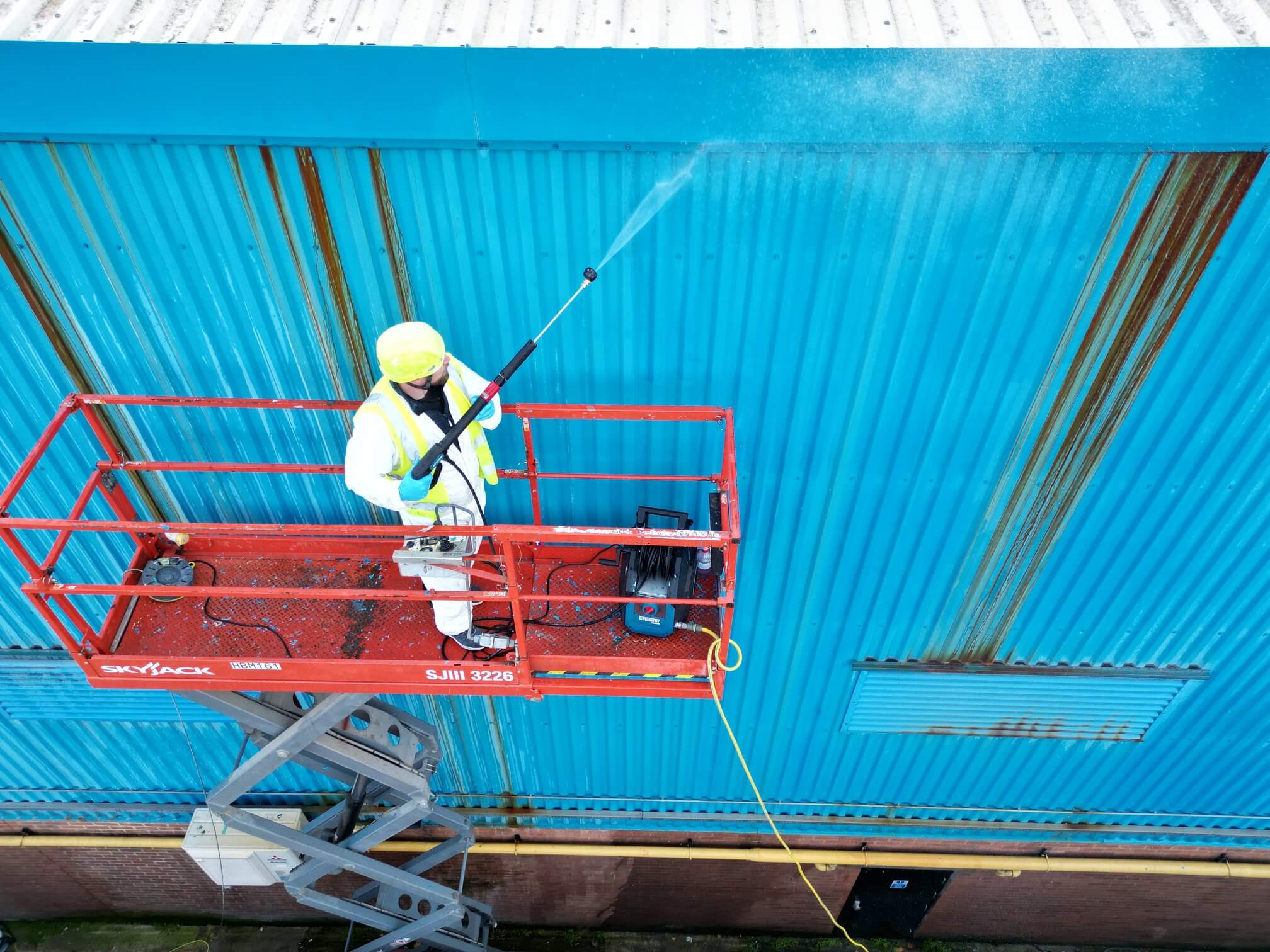 Worker in safety gear pressure washing a blue corrugated metal wall from a red scissor lift.