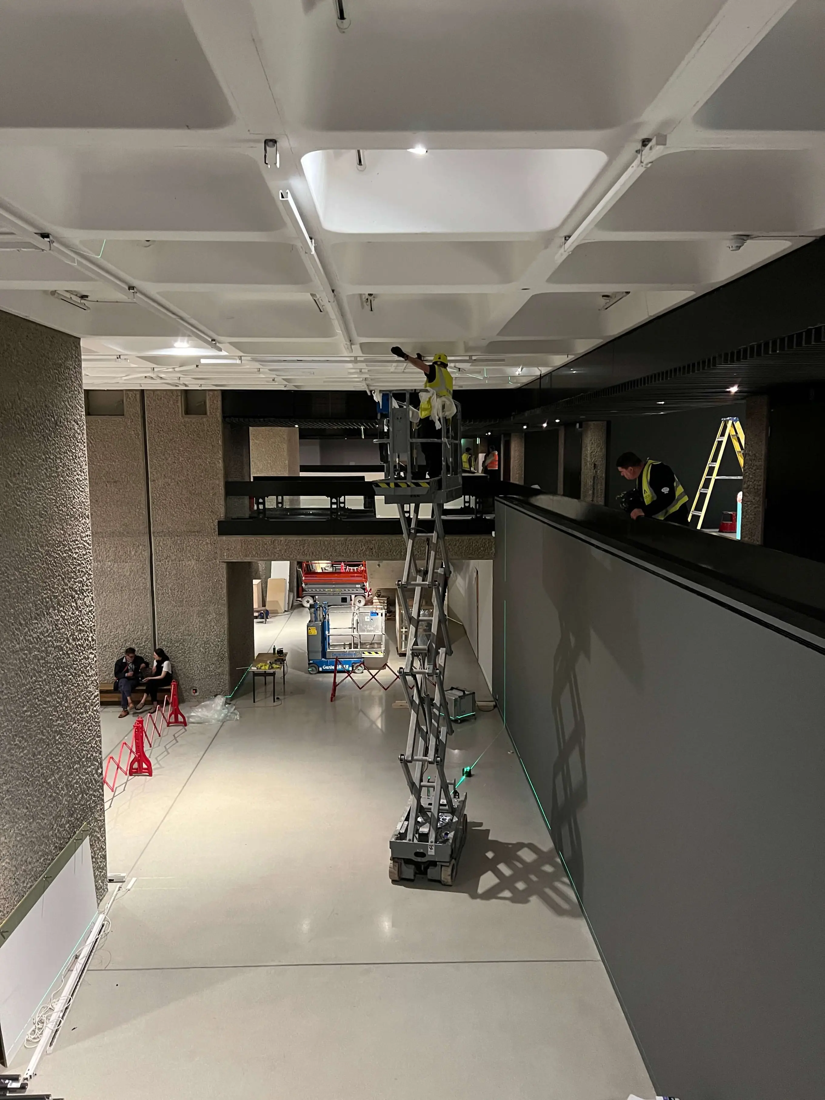 Workers on a raised scissor lift beneath the Barbican Art Gallery ceiling