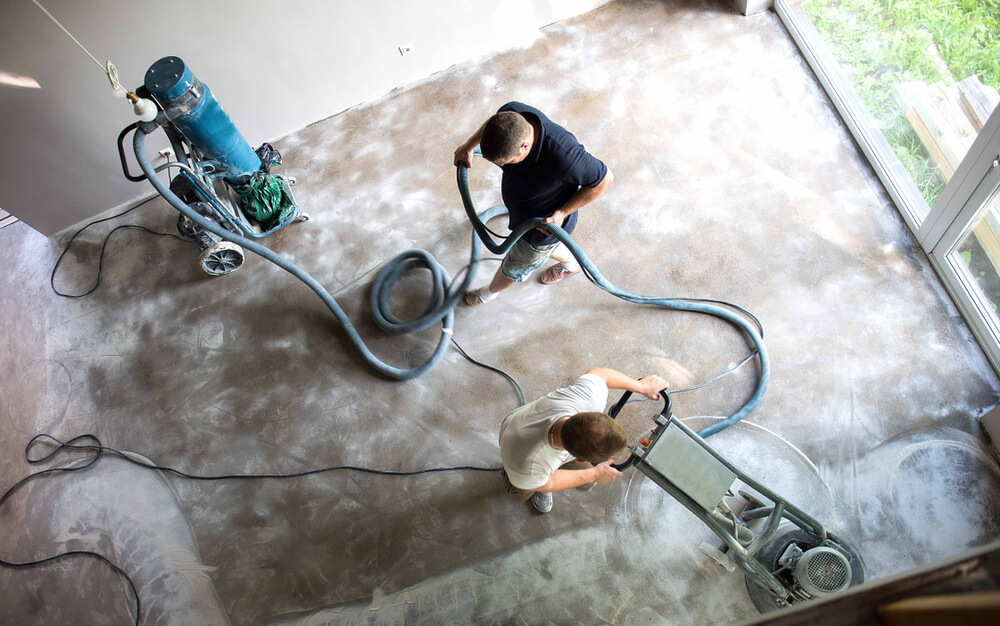 Workers preparing a concrete floor with a floor grinder and dust-extraction equipment.