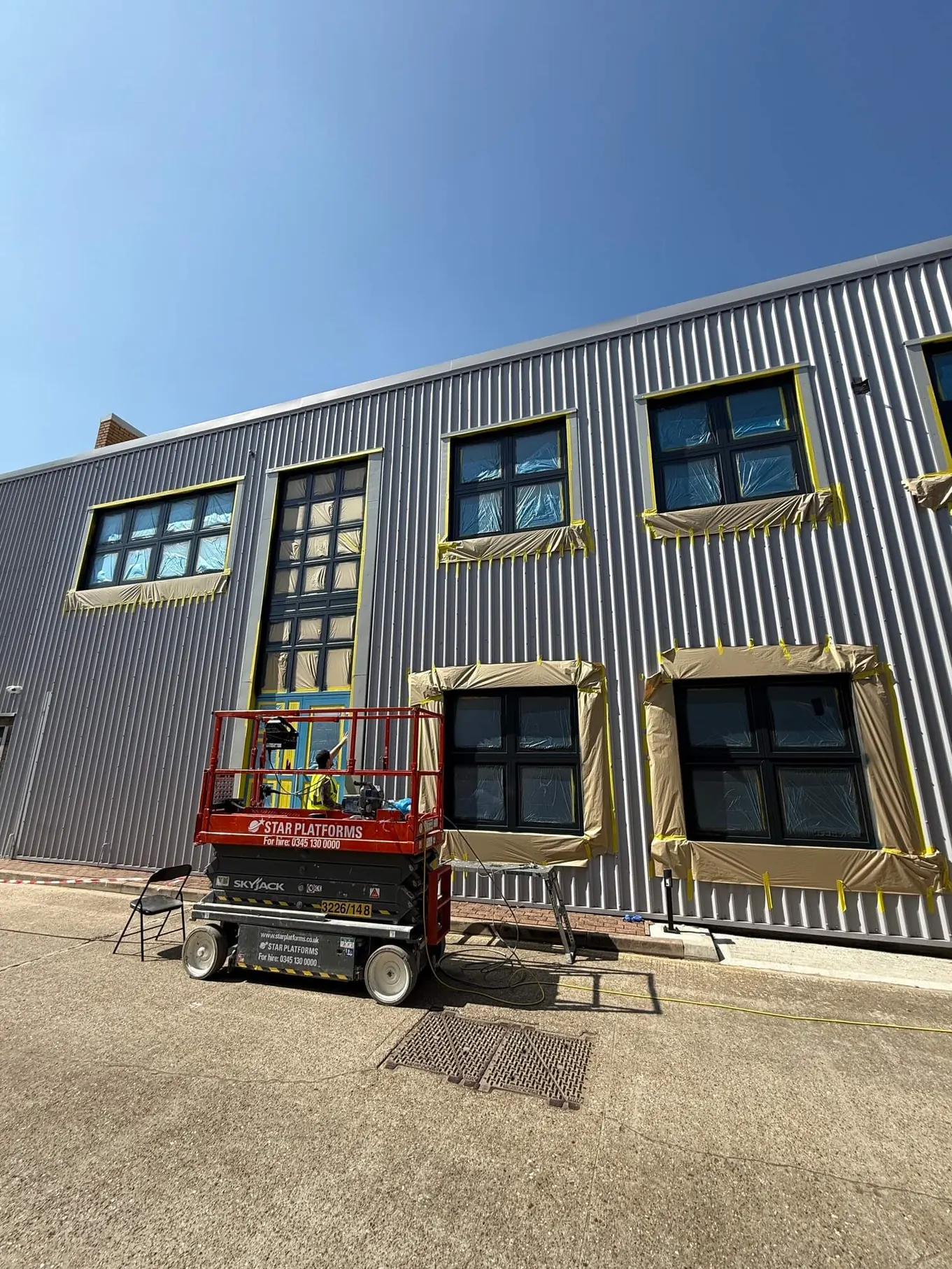 Worker on a red scissor lift preparing a building's windows and door for painting with protective sheeting.
