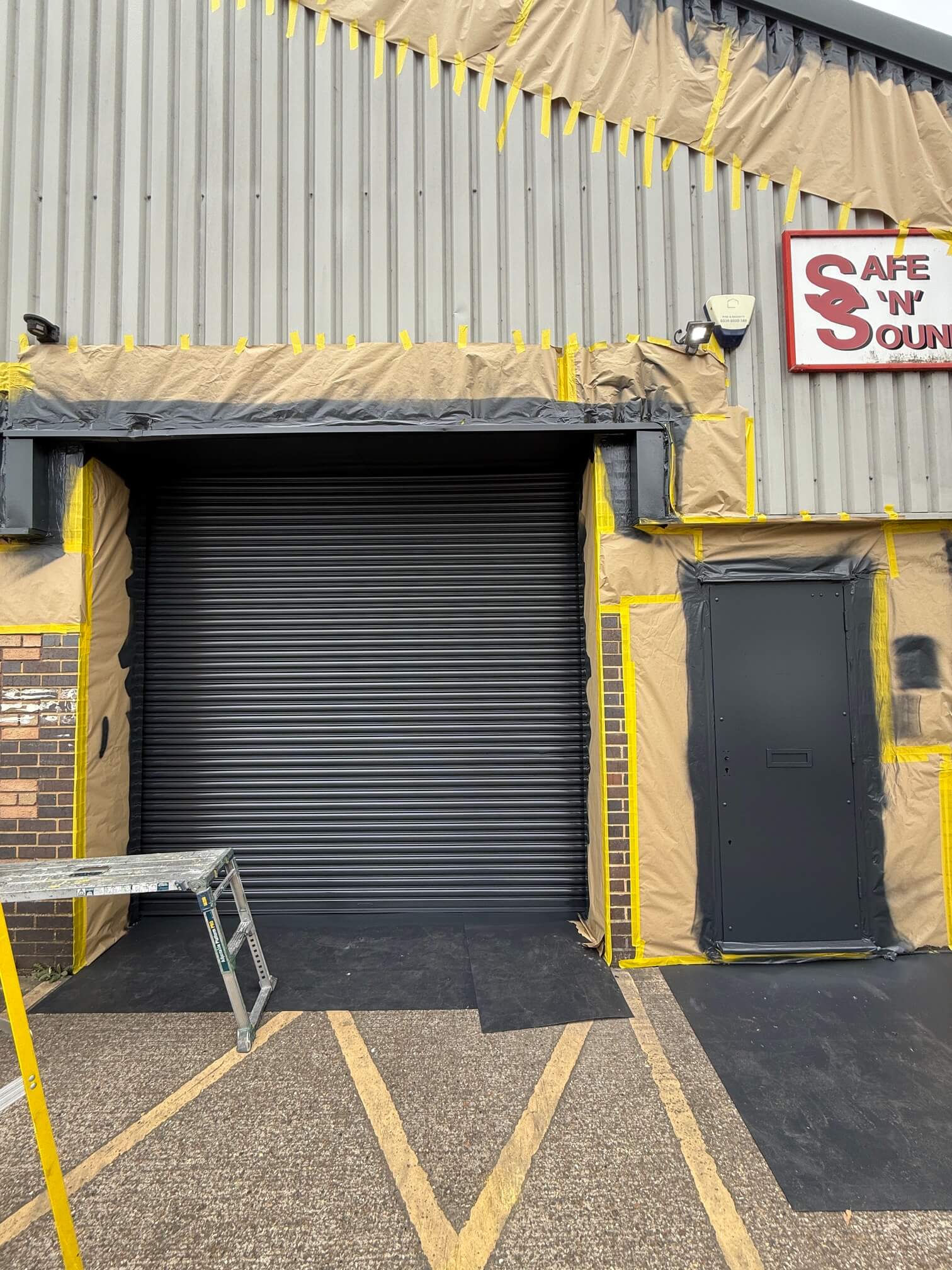 Dark grey industrial roller shutter and adjacent door with protective masking.