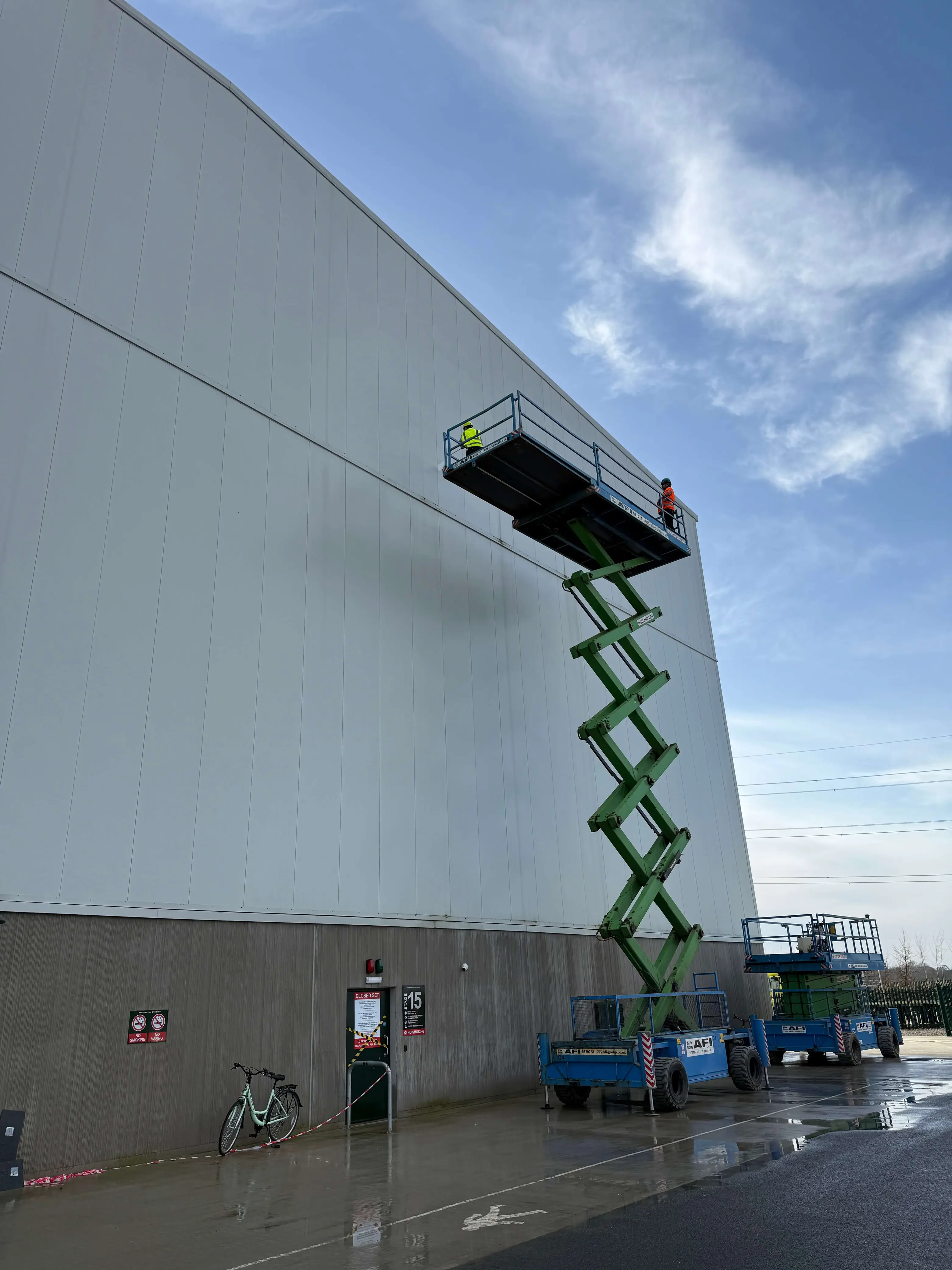 Worker on a raised scissor lift alongside tall light-coloured studio cladding