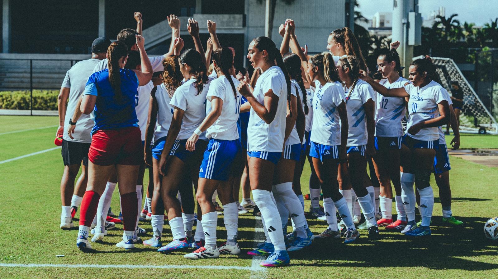 Women’s soccer team huddled in a circle on a grass field raising their hands together.