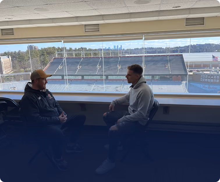 Two men seated facing each other in a room overlooking a stadium with empty bleachers.