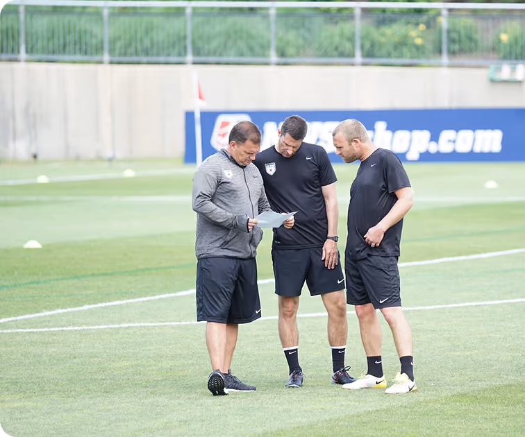 Three men in sportswear standing and looking at a paper on a soccer field.