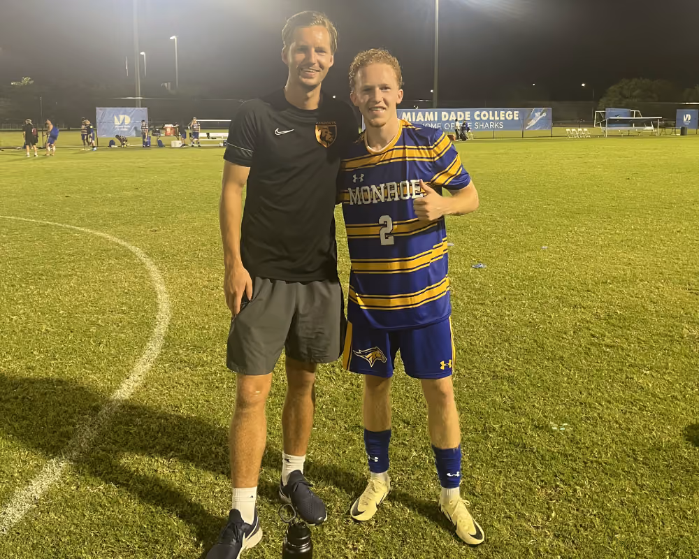 Two male soccer players standing on a lit field at night, one in a Monroe jersey giving a thumbs-up.