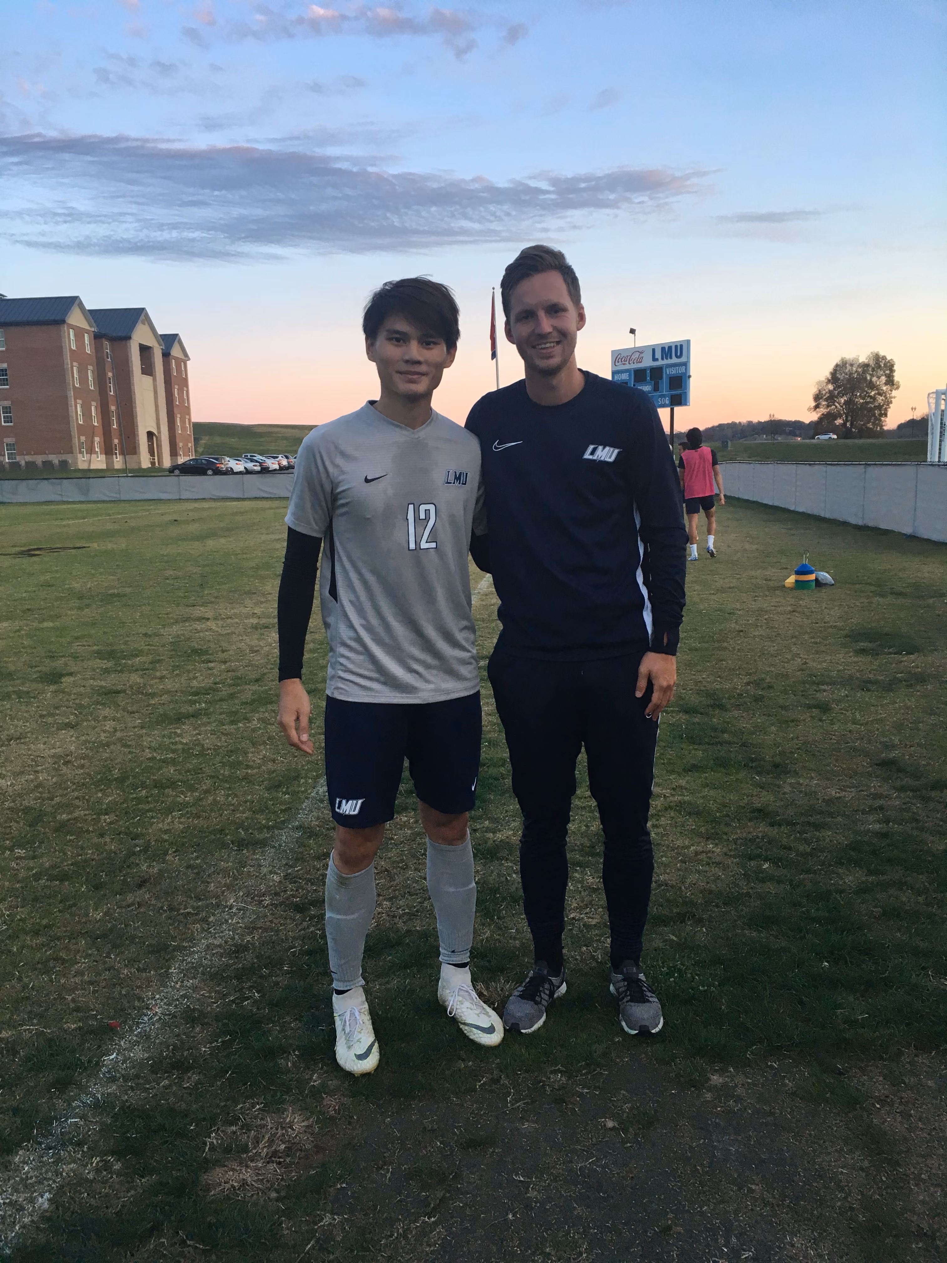 Two men standing on a soccer field at sunset, wearing LMU sports gear, smiling at the camera.