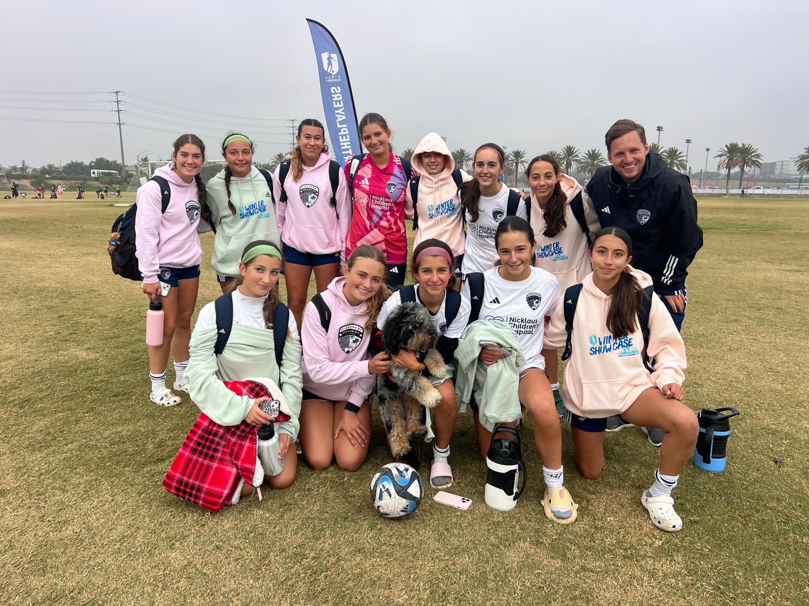 Youth girls' soccer team posing on field with coach, soccer ball, and a small dog during overcast day.