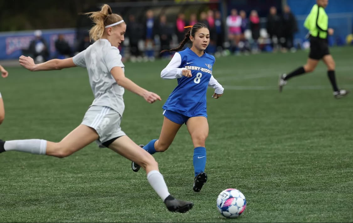 Female soccer player in white kicking ball as opponent in blue runs toward it on green field.