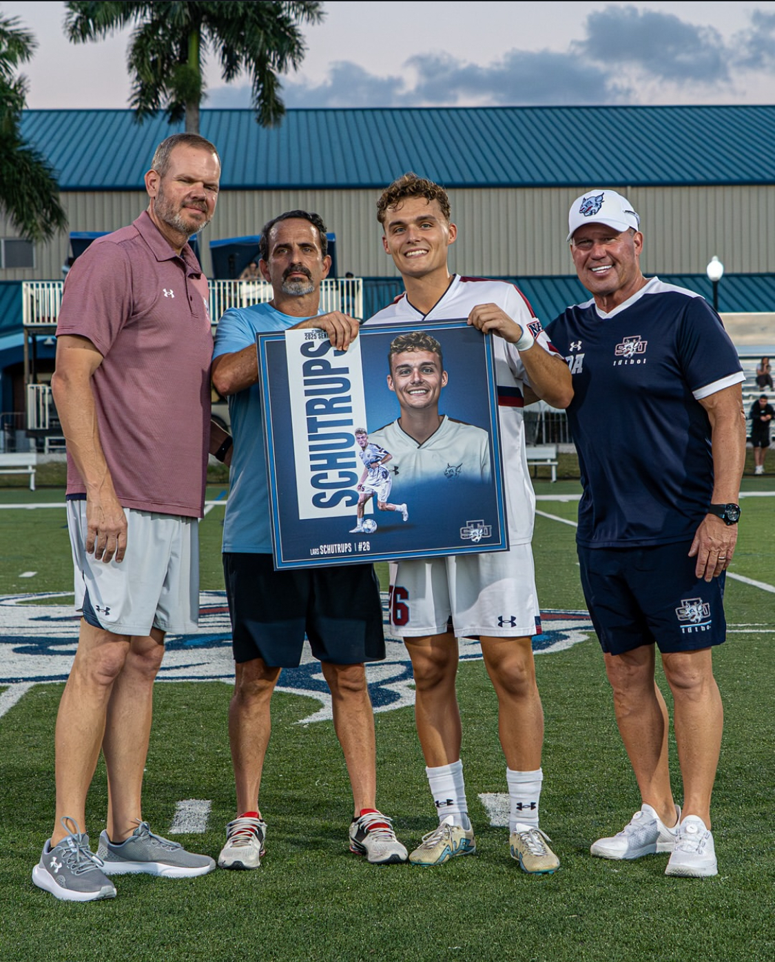 Four men pose on a soccer field, one holding a large poster of a player named Schutrups.