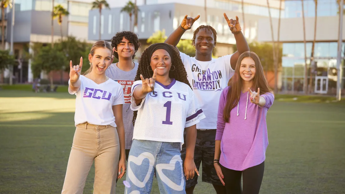 Five diverse students wearing GCU apparel standing outdoors on grass smiling and making hand signs.