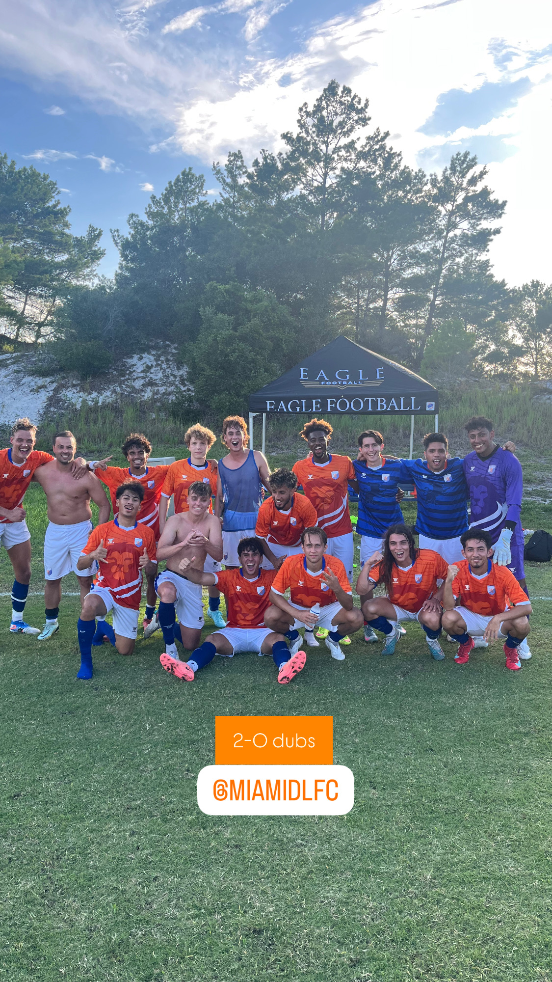 Soccer team in orange and blue jerseys posing on grass near Eagle Football tent after a win.