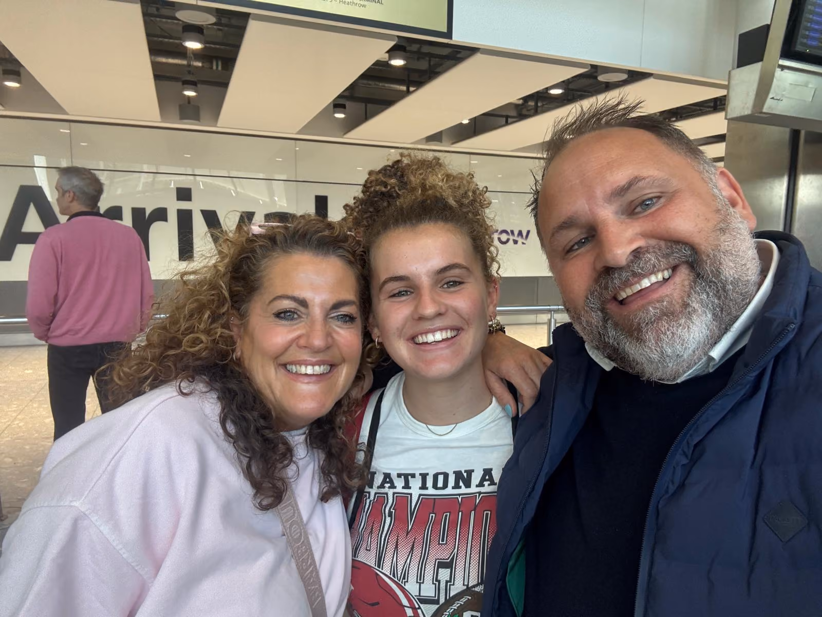 Three people smiling for a selfie at an airport arrivals area with a man in a pink sweater in background.