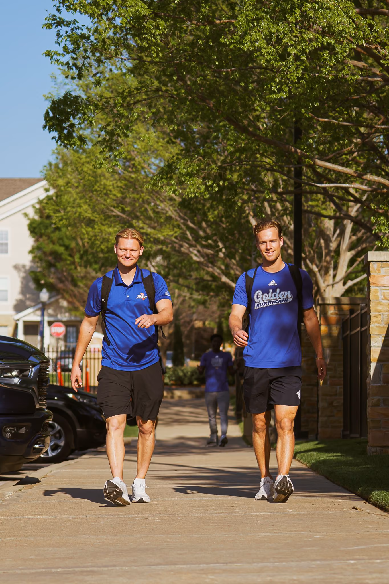 Two young men wearing blue shirts and backpacks walking on a sunny tree-lined sidewalk.