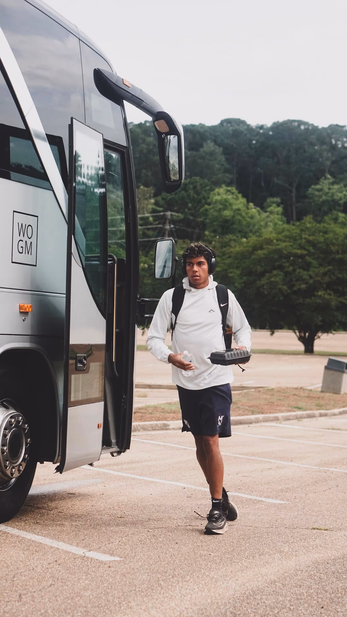 Young man with backpack, headphones, drink, and food walking from a parked tour bus.