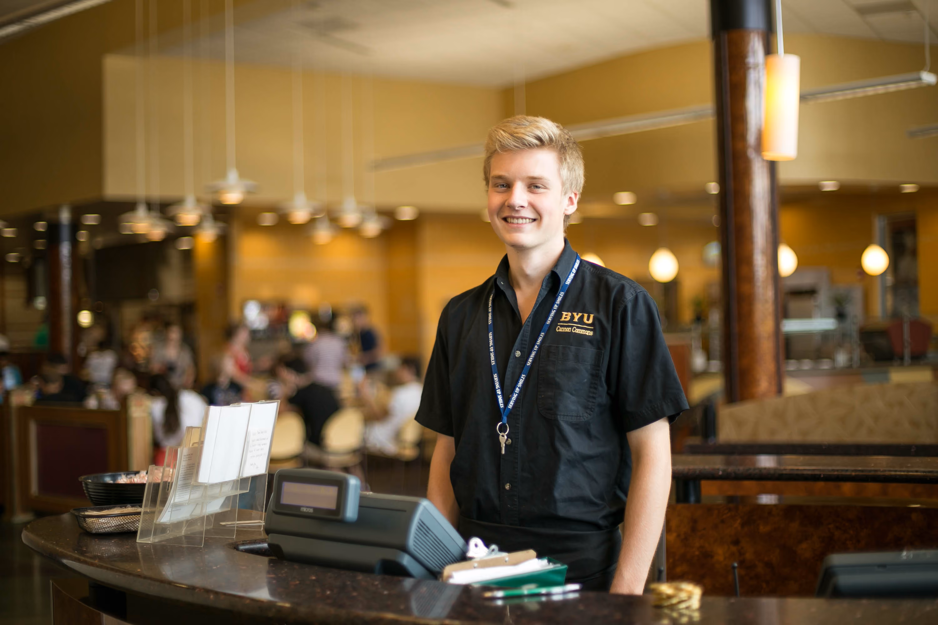 Smiling young man in BYU Canon Commons uniform standing behind a counter with a register.