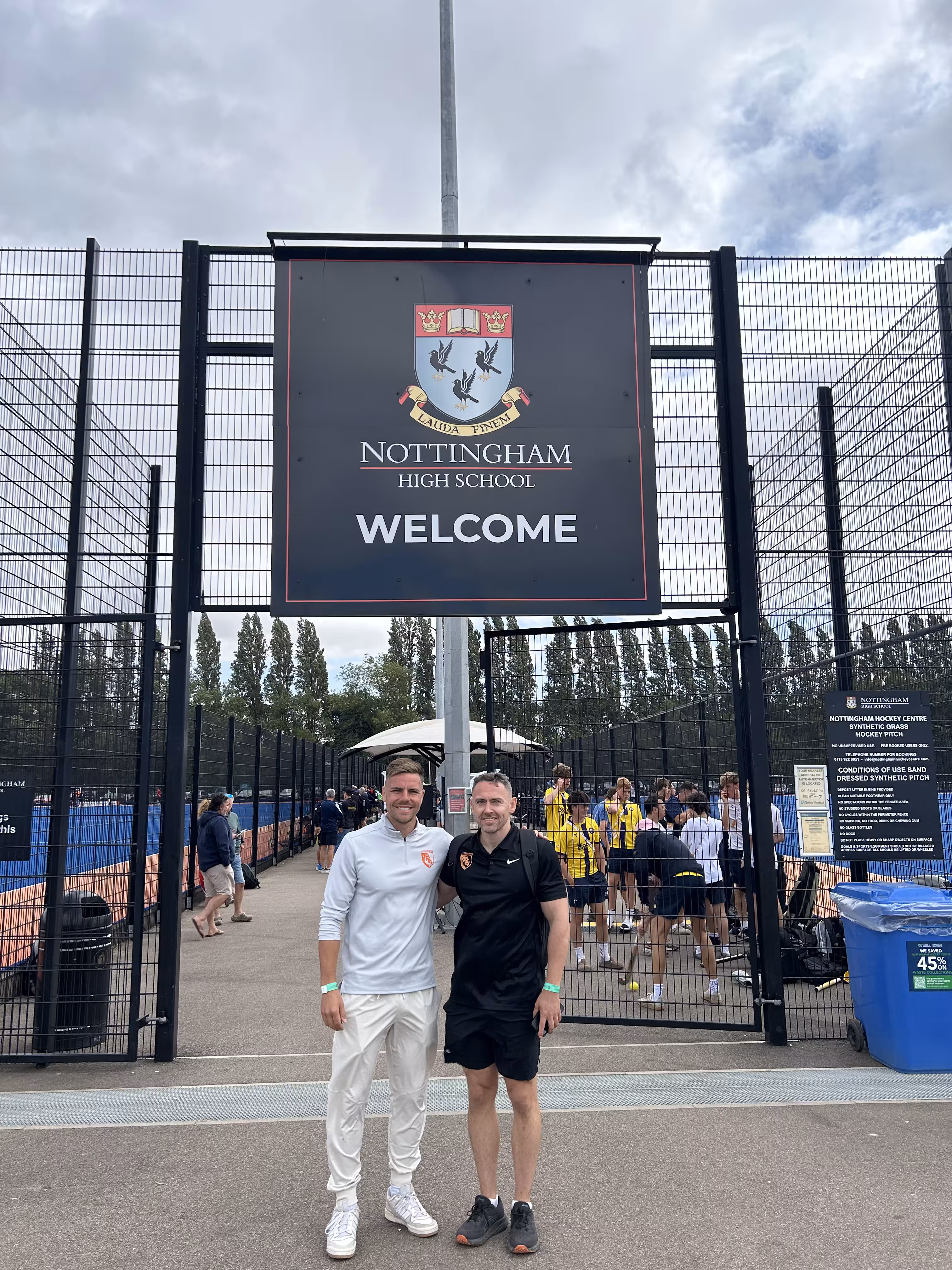 Two men standing in front of Nottingham High School welcome sign by a sports pitch with players in the background.