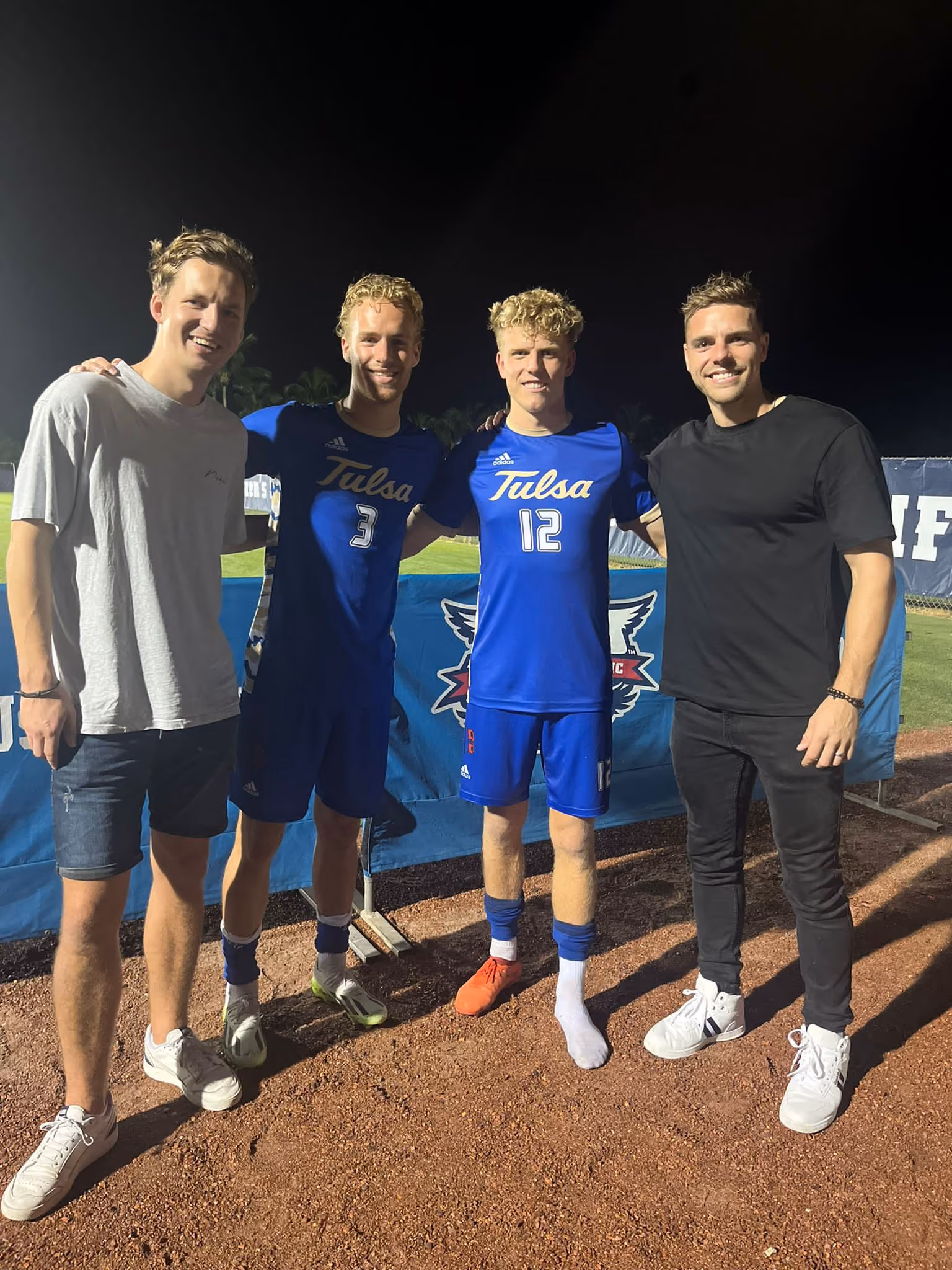 Four men smiling at night, two in Tulsa soccer uniforms, two in casual clothes on a sports field.