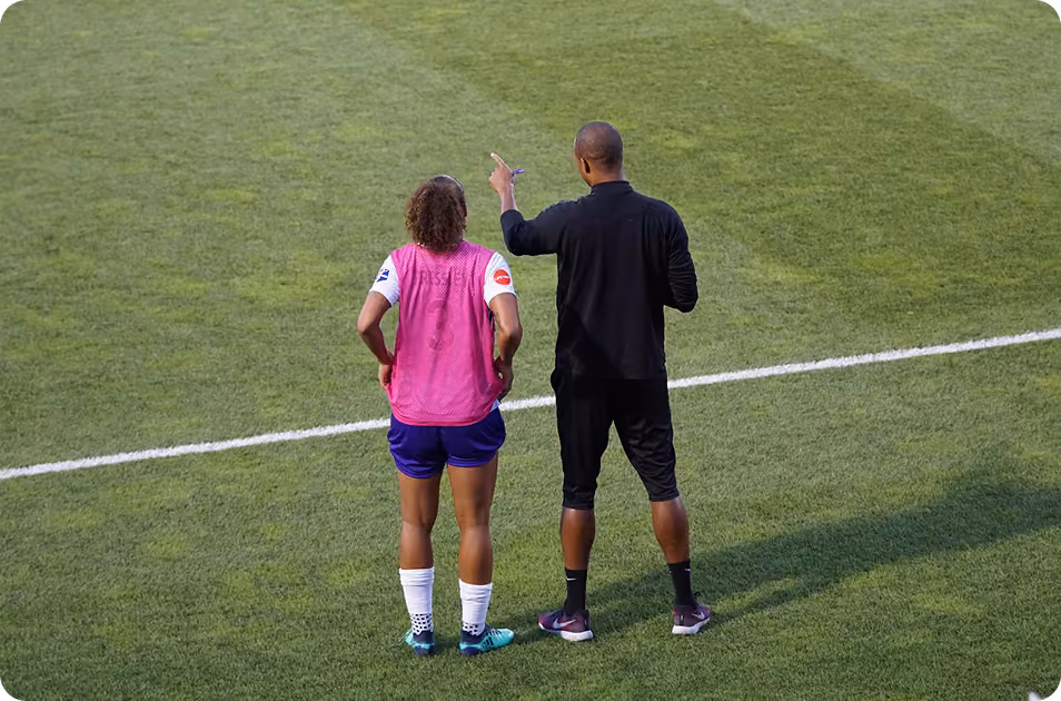 Coach in black talks to a soccer player in pink vest on green field near white line.